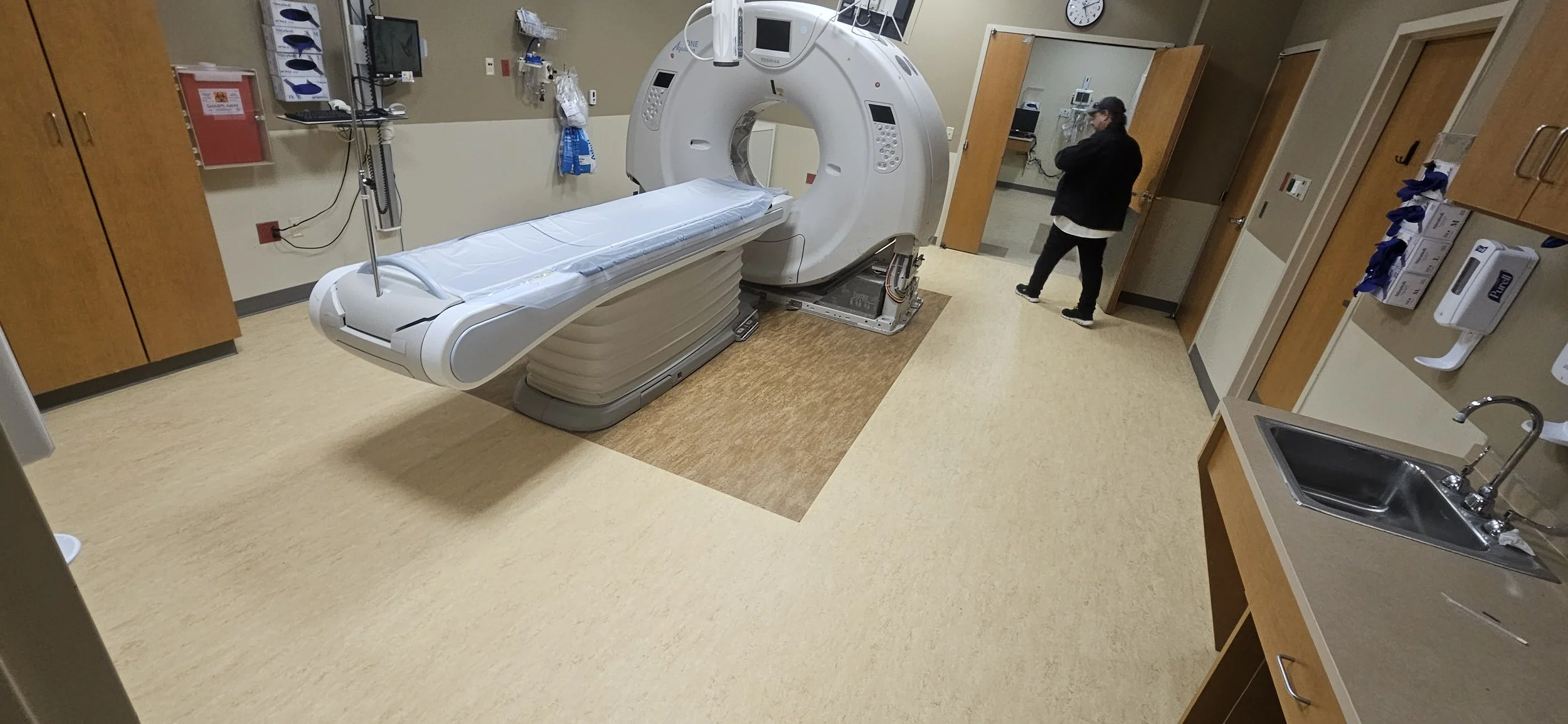 A hospital room with a large MRI machine, a patient bed, medical equipment, cabinets, sink, and a person standing near the door.