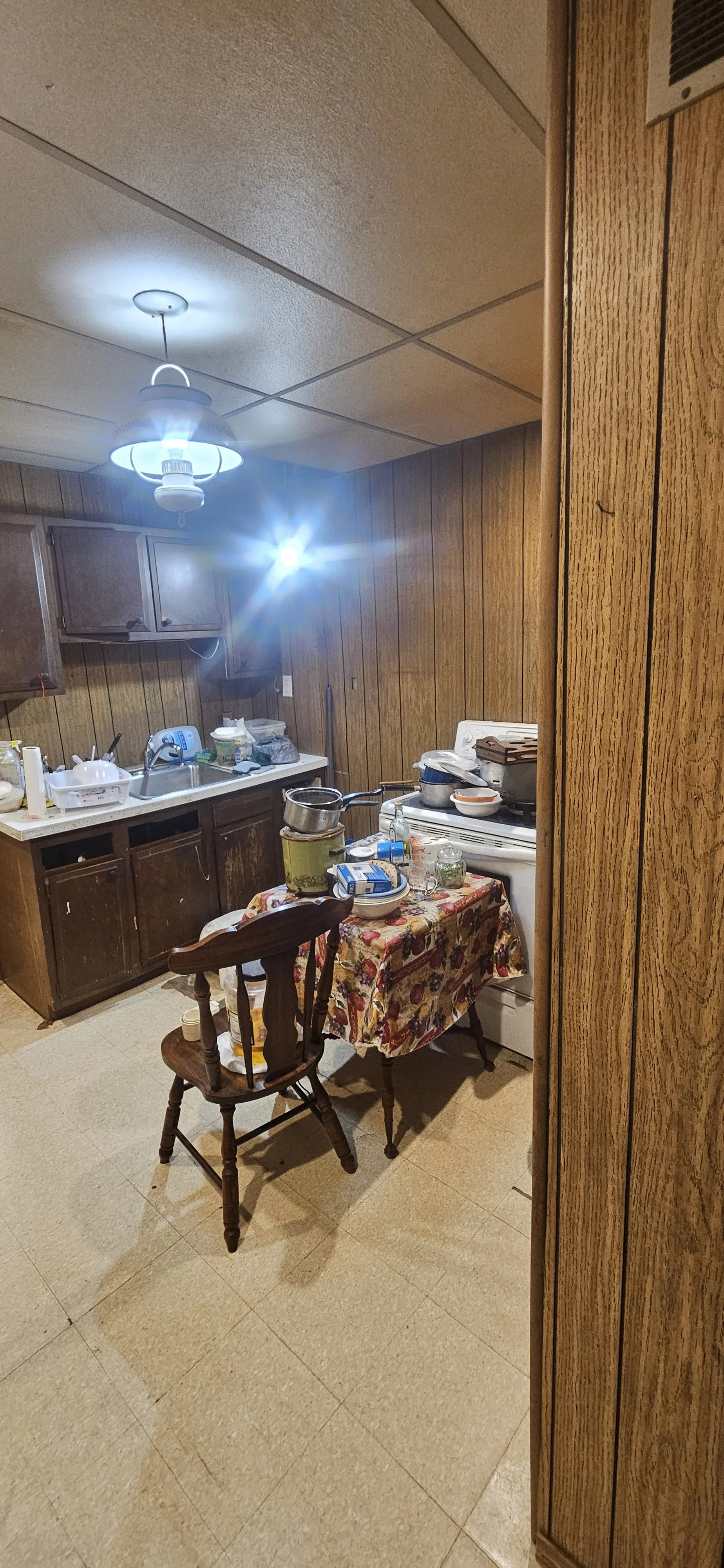 A cluttered kitchen with wooden paneling, a small dining table with a floral tablecloth, and various kitchen items on the counter and table.