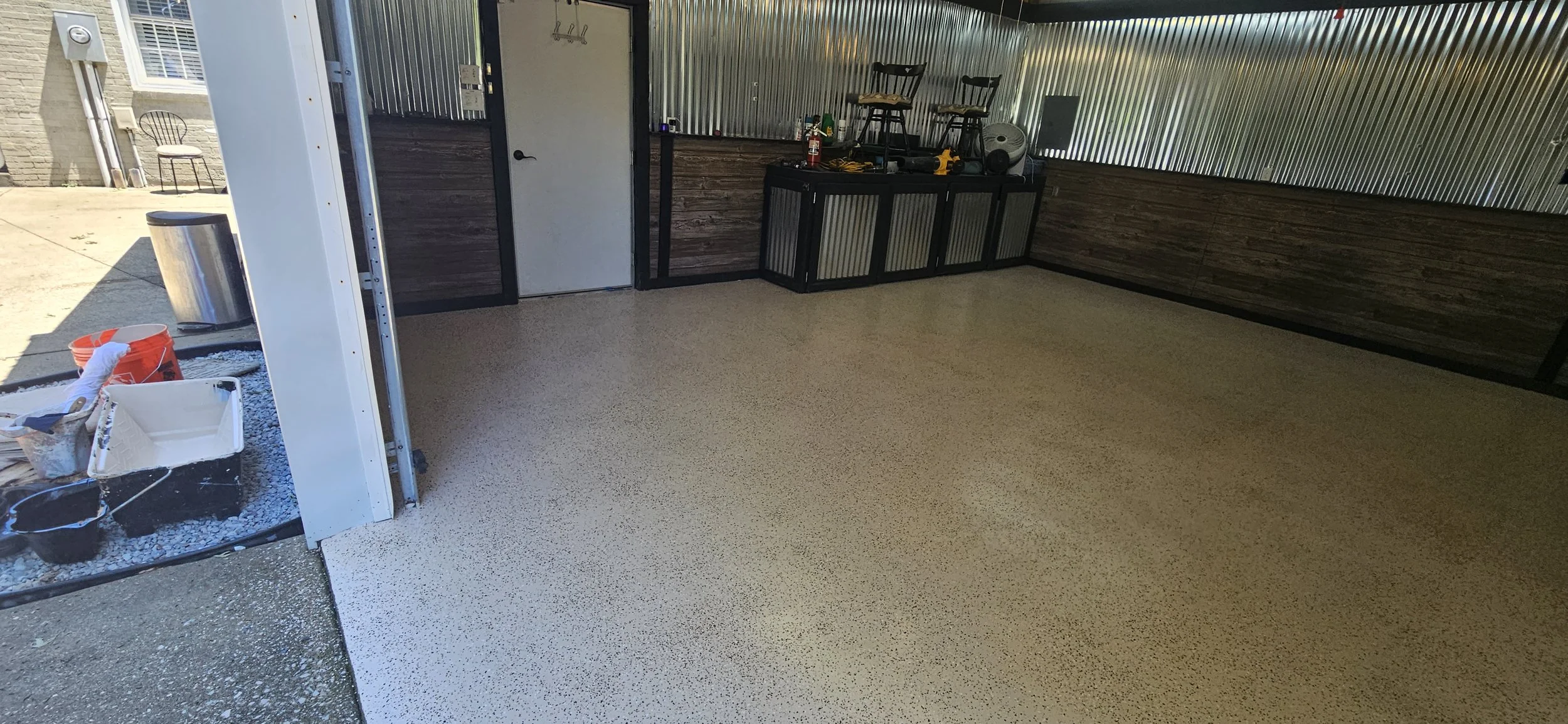 Photo of a partially finished garage or workshop with a speckled epoxy floor, wooden walls, and a corrugated metal ceiling. The area contains some tools and chairs on a black cabinet, with a door leading into the interior. Outside, there's a bucket, 