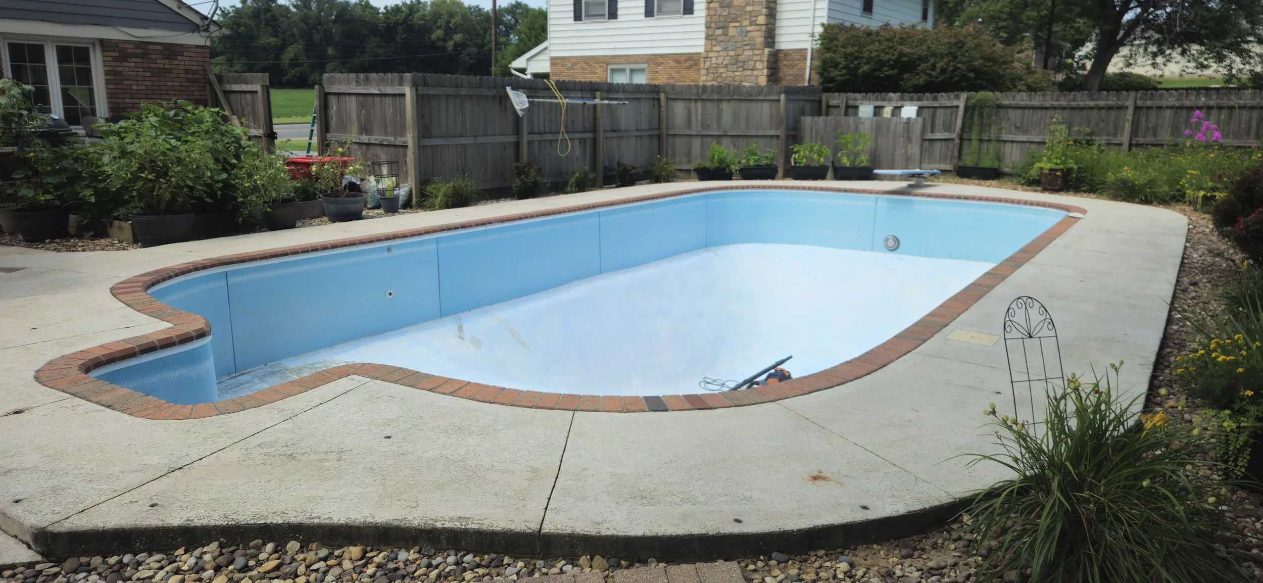 An empty kidney-shaped swimming pool with a brick border surrounds the interior blue pool liner. The pool is located in a backyard with a concrete deck, various potted plants around the perimeter, and a wooden fence enclosing the yard. There are some