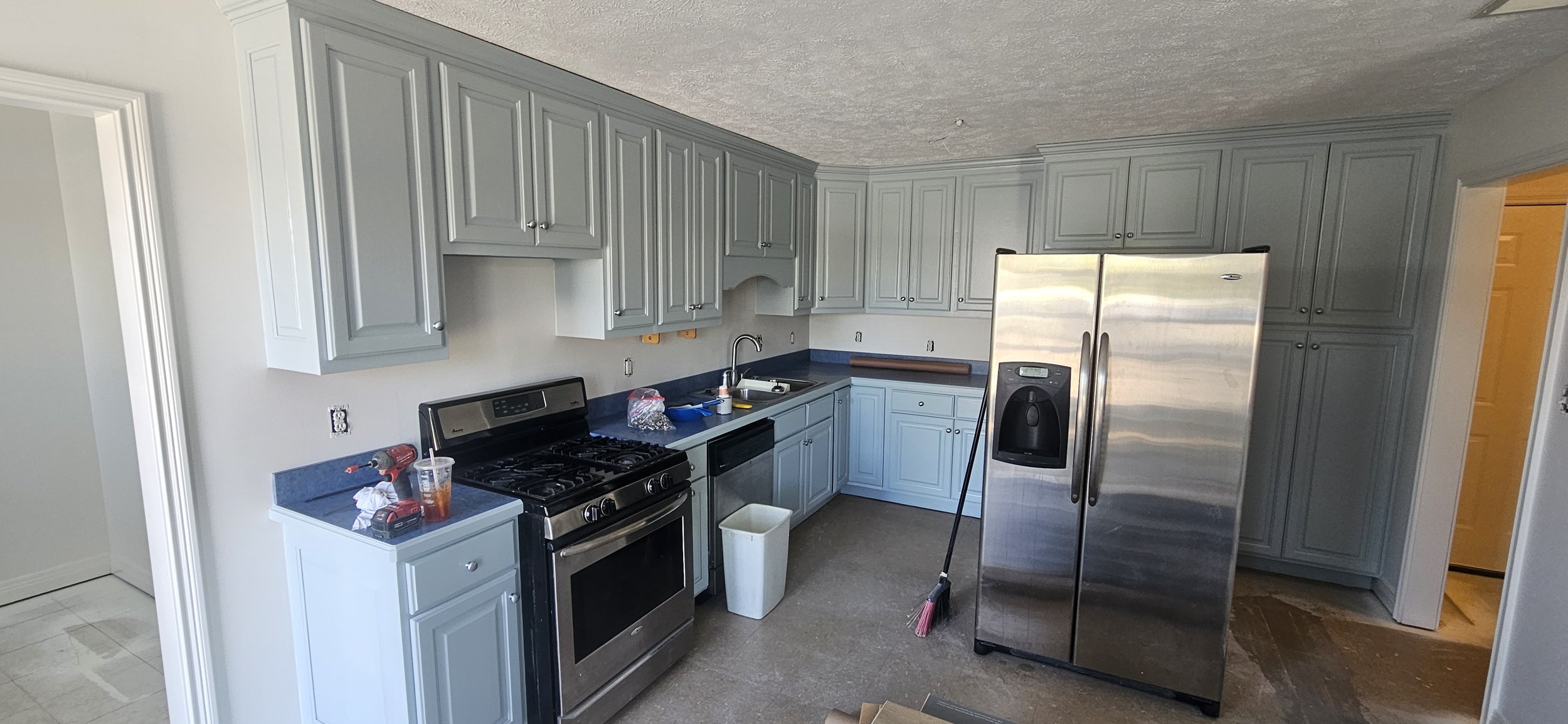 A kitchen with light blue cabinets, stainless steel appliances including a refrigerator, stove, and dishwasher, and a black countertop. There are some tools, a drink, and cleaning supplies on the counter, and a mop leaning against the refrigerator.