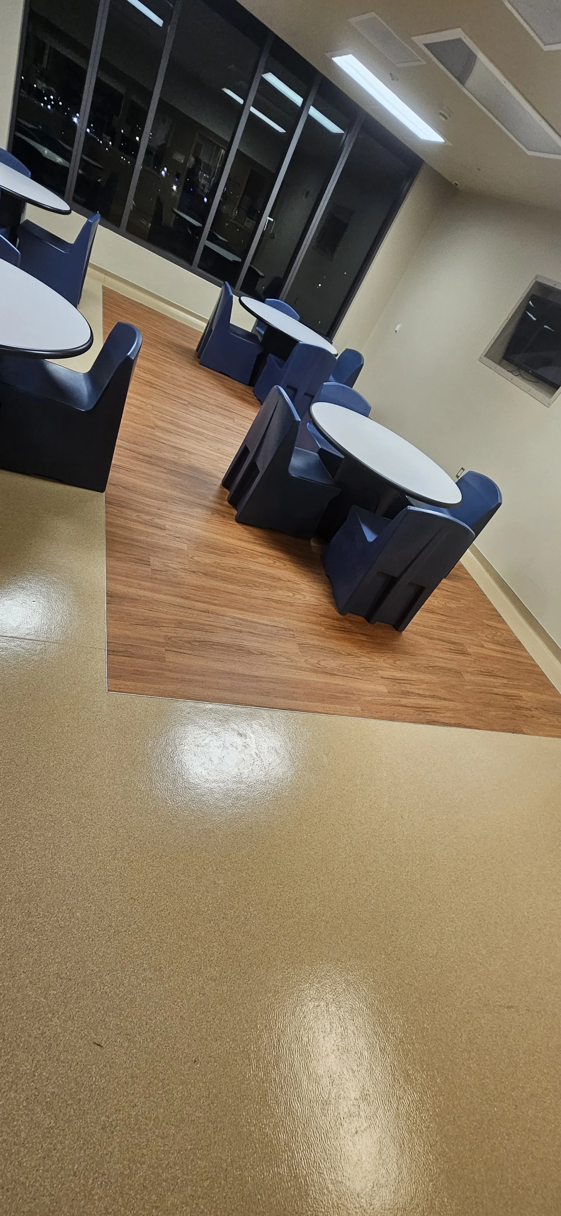Empty dining area with round tables, blue chairs, and hardwood flooring section in a modern room at night.
