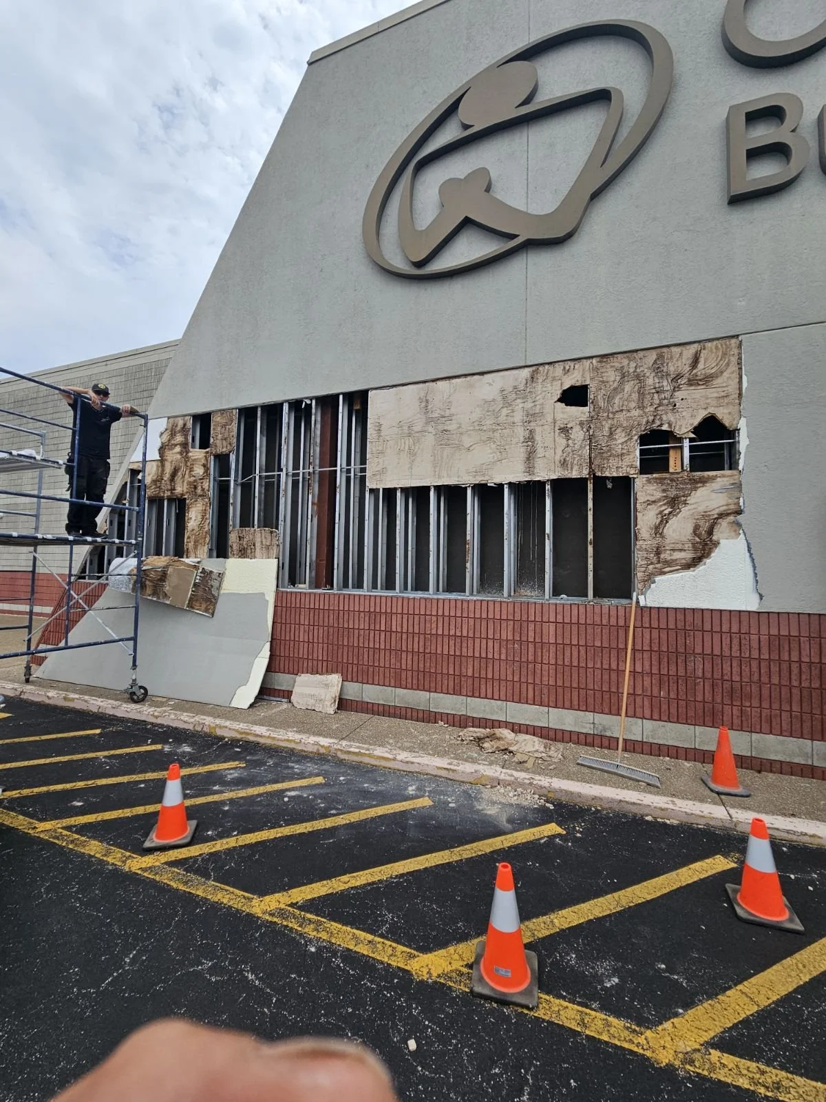 A worker on scaffolding repairs the damaged exterior wall of a building, which has exposed wooden and metal framing and boarded-up sections. Orange traffic cones are placed on the parking lot in front.