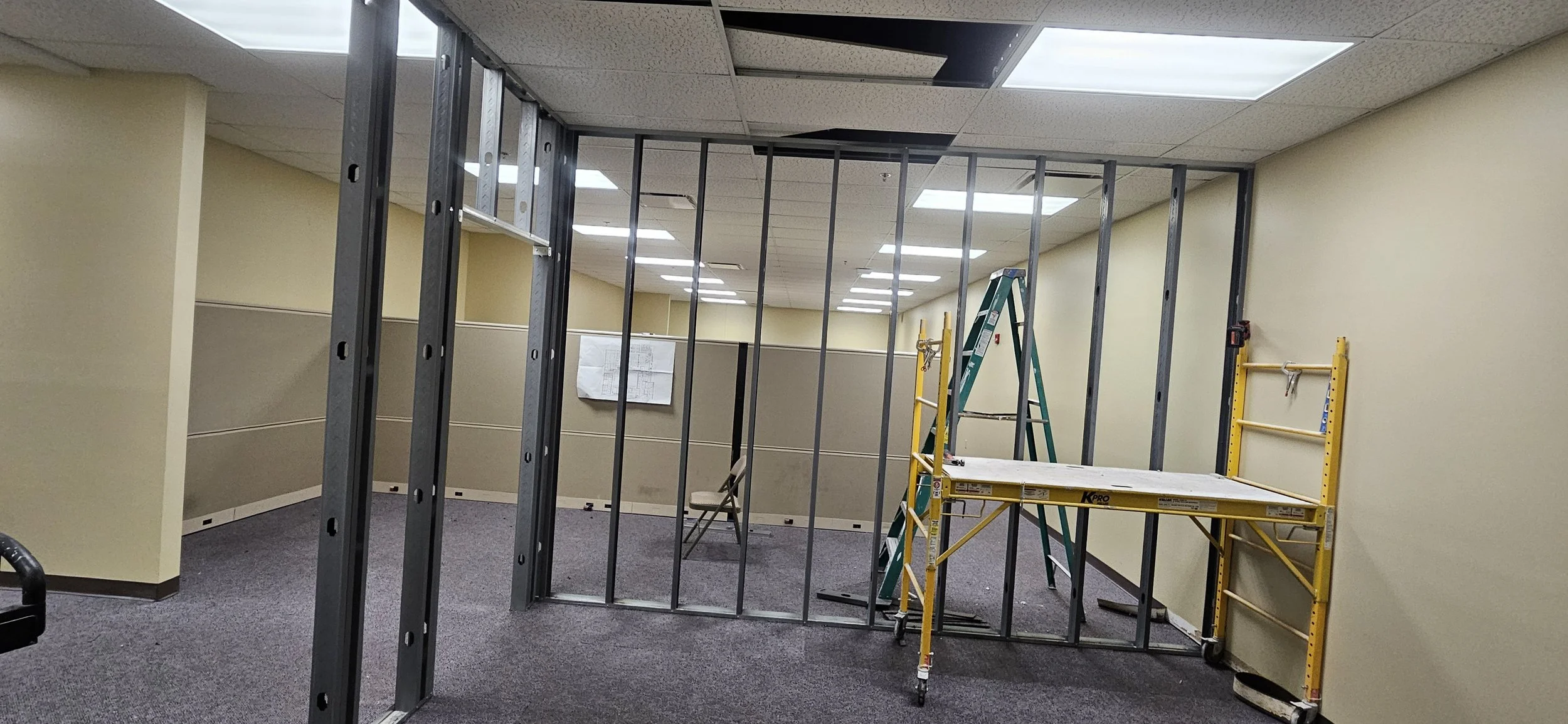 Office renovation with metal wall framing, a green ladder, a yellow scaffold, and construction papers on the wall.