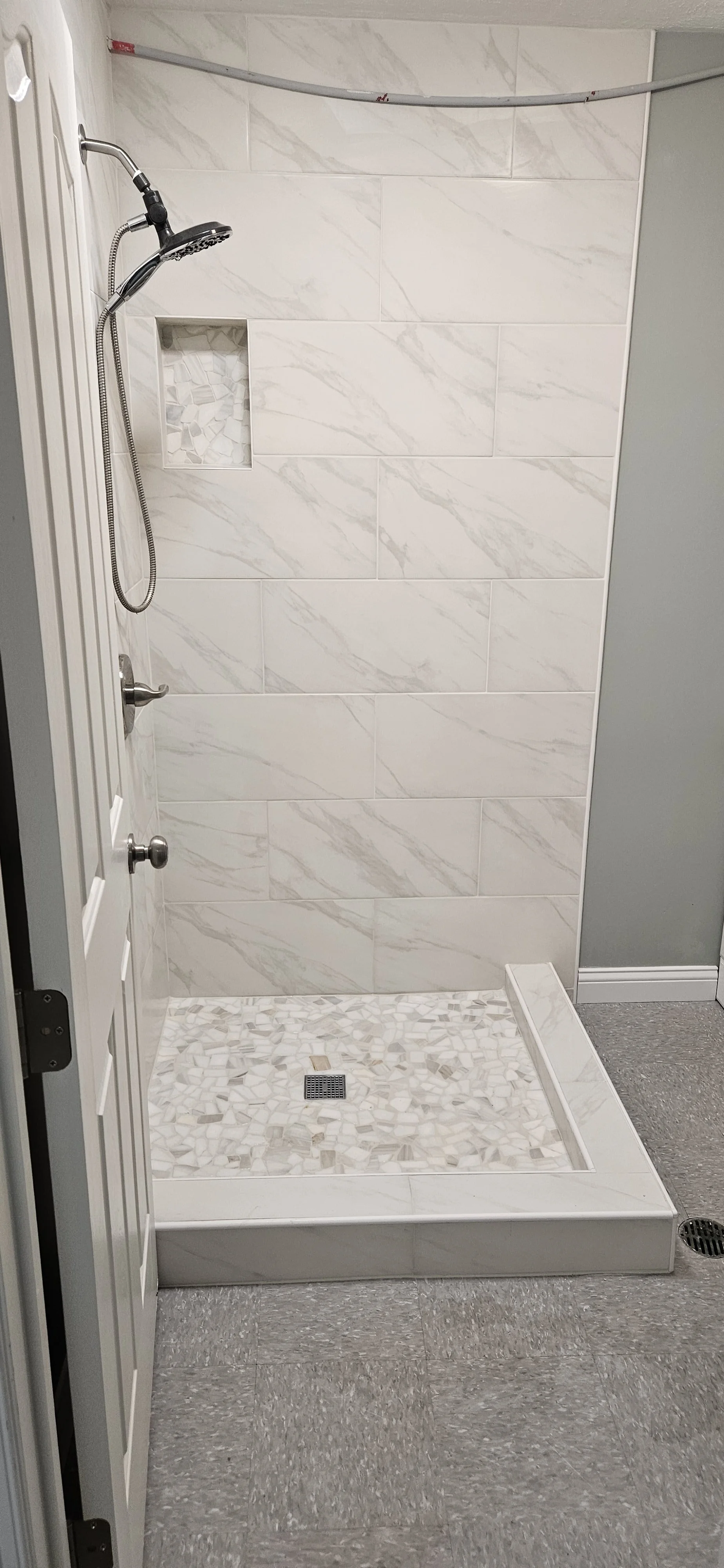 A walk-in shower with light-colored marble tiles on the walls, a mosaic tile floor, a built-in niche with mosaic stones, a handheld showerhead, and a square drain in the floor. The shower is enclosed with a partial door and adjacent to a gray wall an
