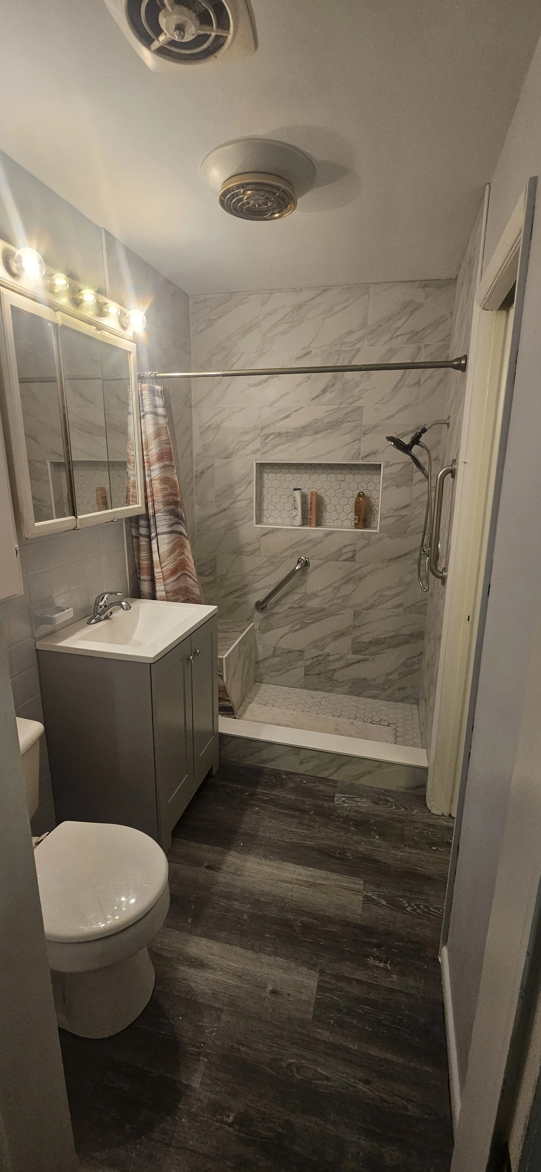 Small bathroom with a grey vanity, white sink, toilet, and a walk-in shower with marble-tile walls, a built-in niche with toiletries, a showerhead, a grab bar, and a curtain on a curtain rod. Dark wood flooring and a ceiling with a smoke detector and