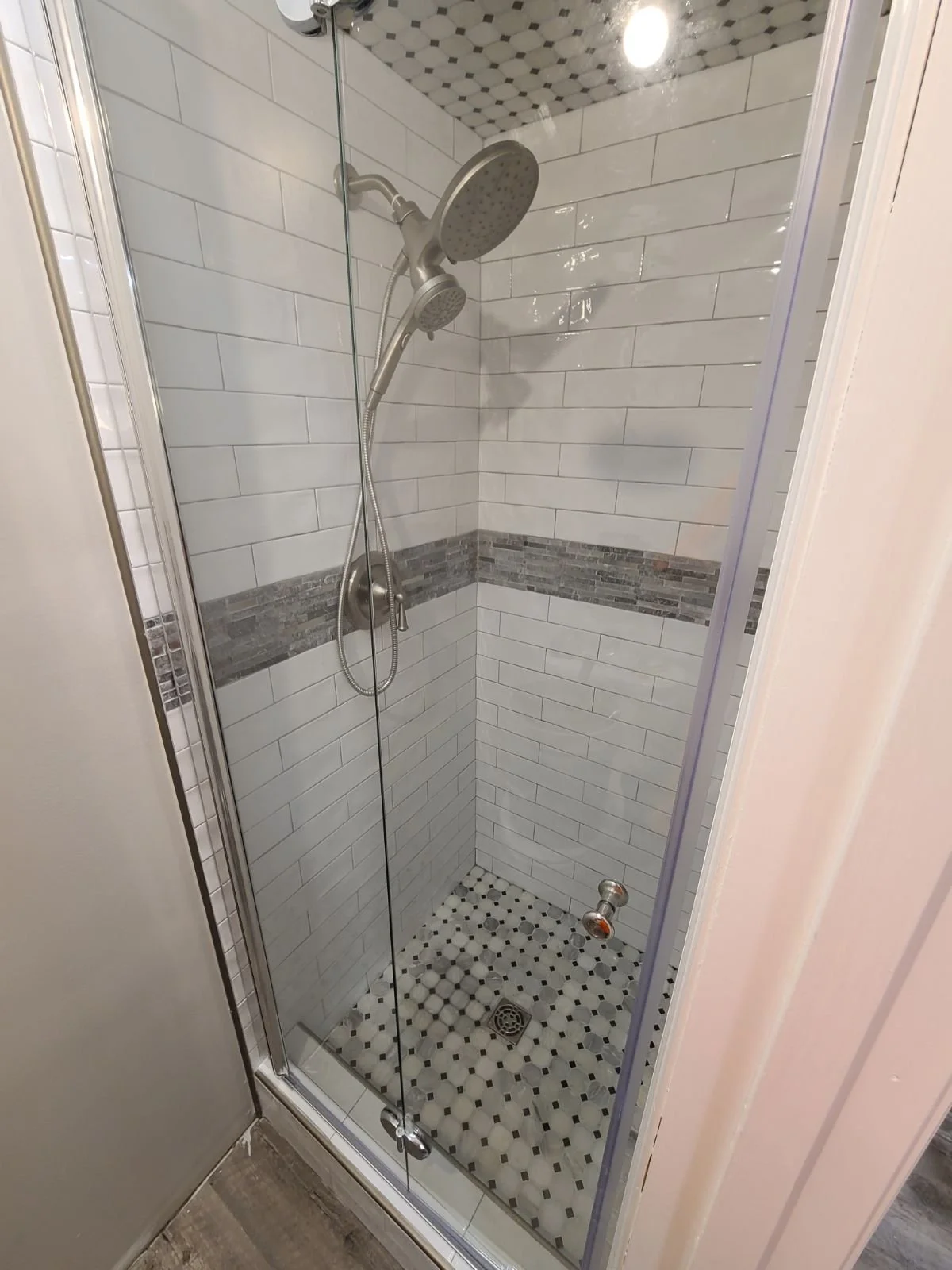 A corner shower with white subway tile walls, a horizontal band of gray mosaic tile, and black and white hexagon floor tiles. Contains a dual showerhead and a glass door.