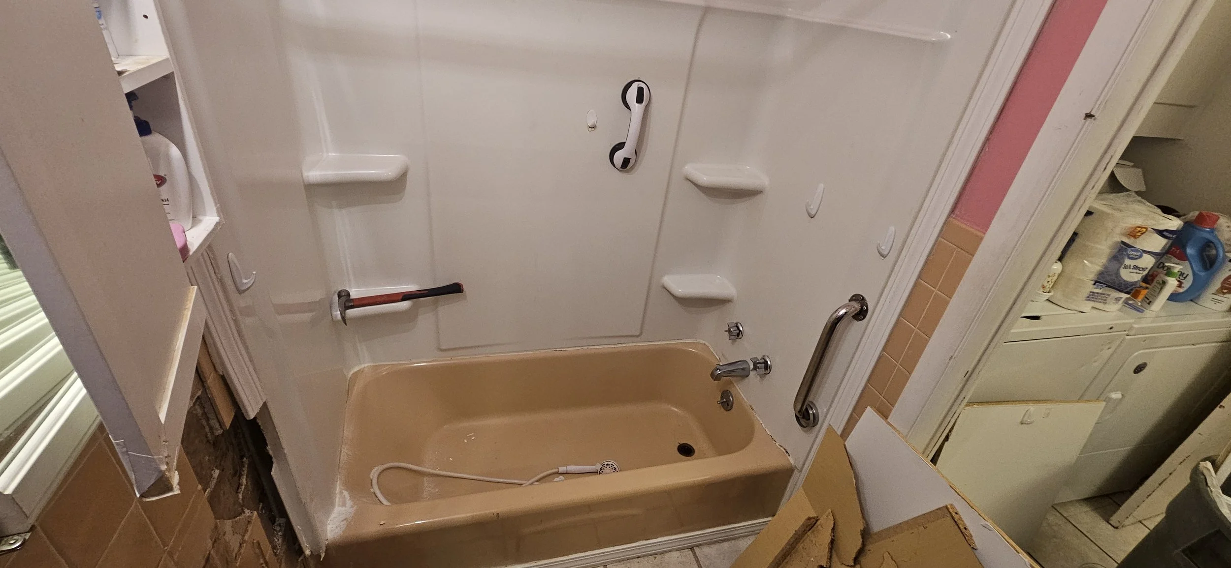 Bathroom with a bathtub, grab bars, soap shelves, and a showerhead. The bathtub is beige, and the walls have various personal care items, with some tile and wall damage visible.