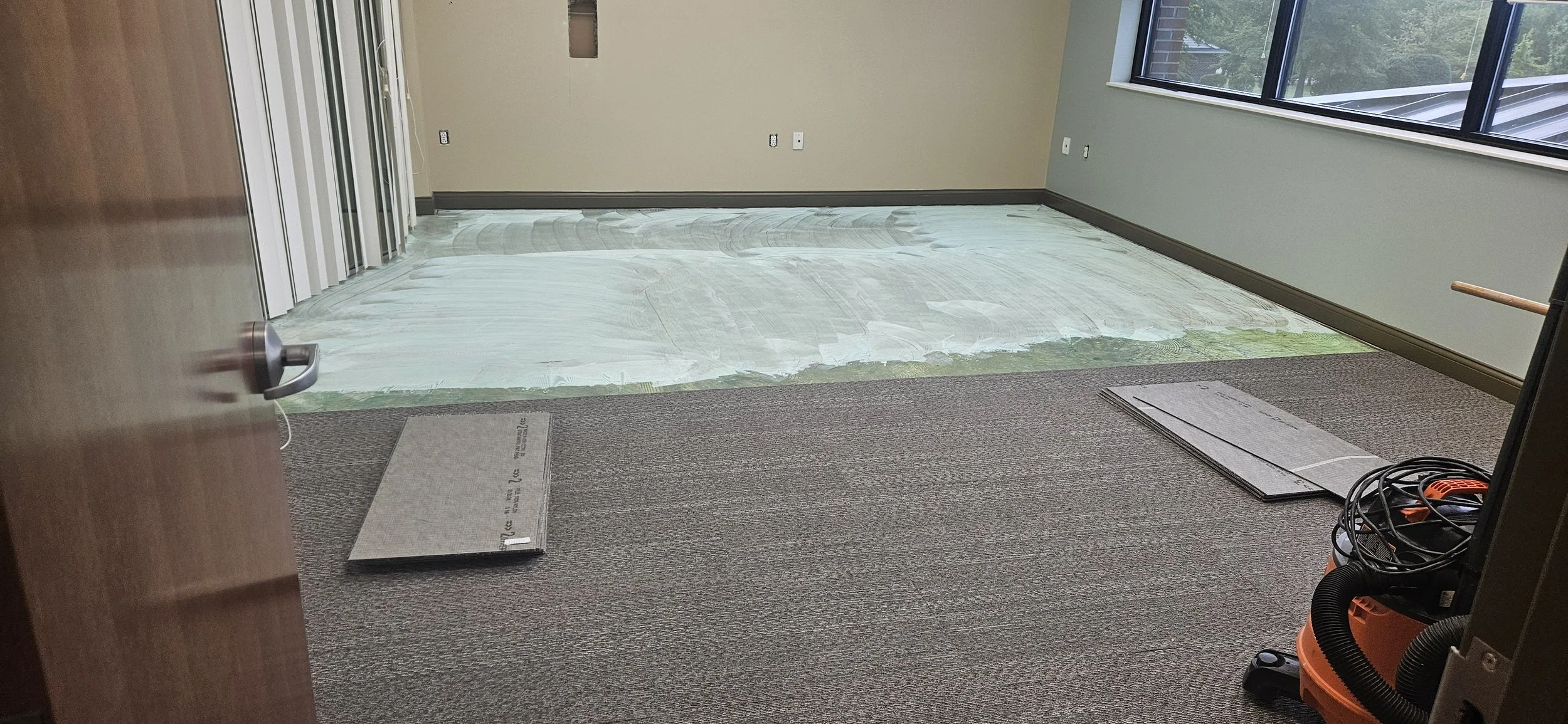 Office room under renovation with a protective film on the floor, carpeted area, and large windows showing trees outside.