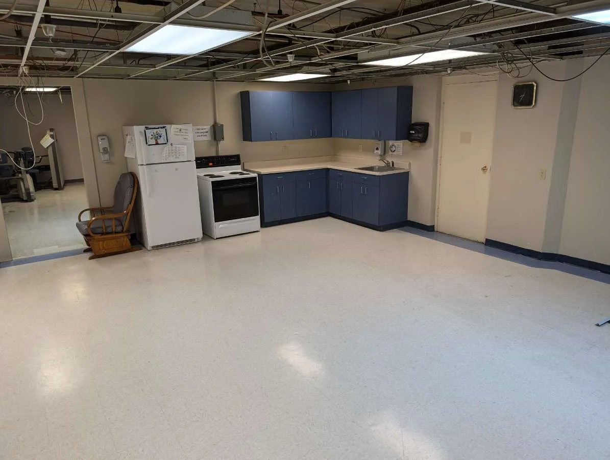 A mostly empty room with a small kitchenette, blue cabinetry, a white refrigerator, stove, and sink. An armchair is placed next to the refrigerator. The ceiling is exposed, showing wiring and lighting fixtures. There's a clock on the wall and a close