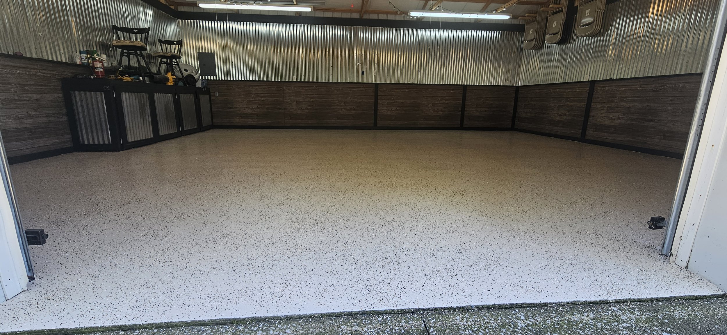 Empty garage with speckled white floor, wood paneling on the lower walls, and silver corrugated metal on the upper walls and ceiling. Tools and chairs are stored on top of black storage cabinets on the left side.
