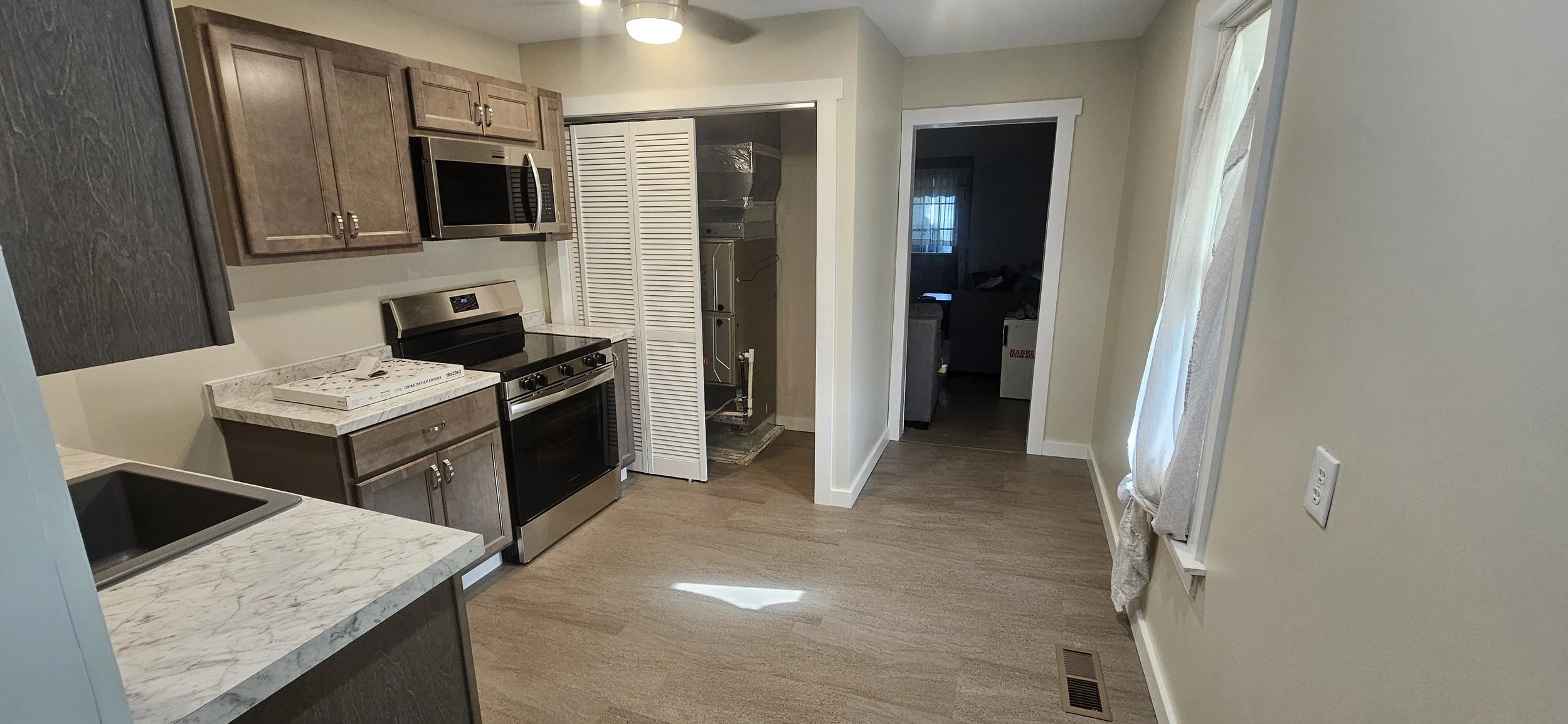 A kitchen with brown cabinets, stainless steel stove, microwave, and marble countertops. There is a small window with curtains, and a visible laundry area with a closet door open and some appliances inside.