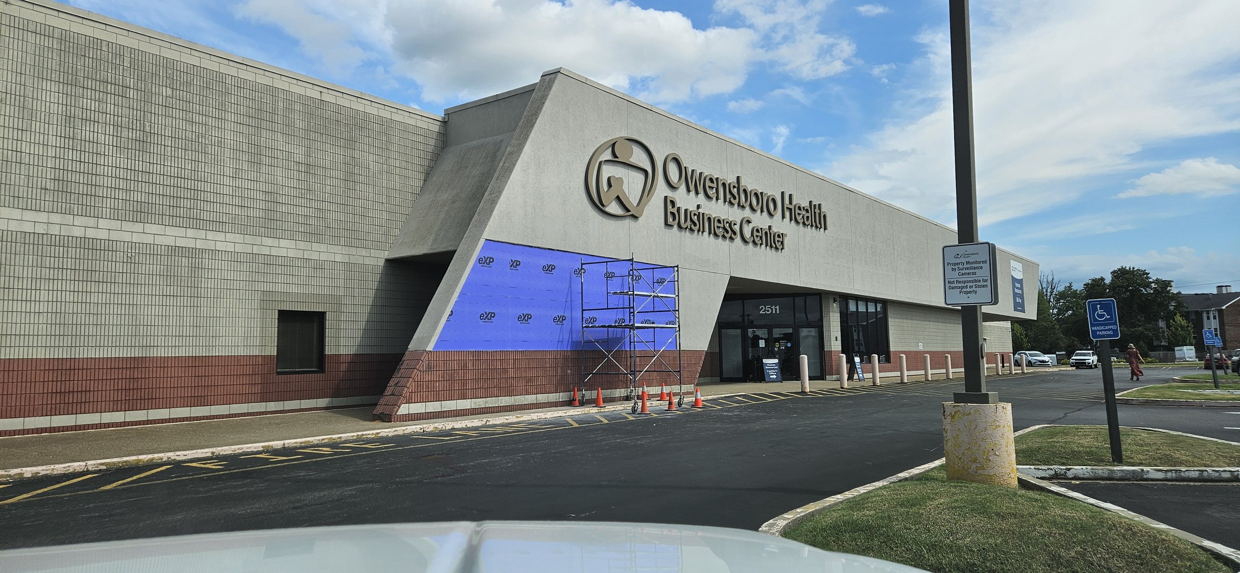 Exterior of Owensburg Health Business Center with scaffolding and construction materials near the entrance, parking lot with reserved disabled spots, and blue sky with some clouds.