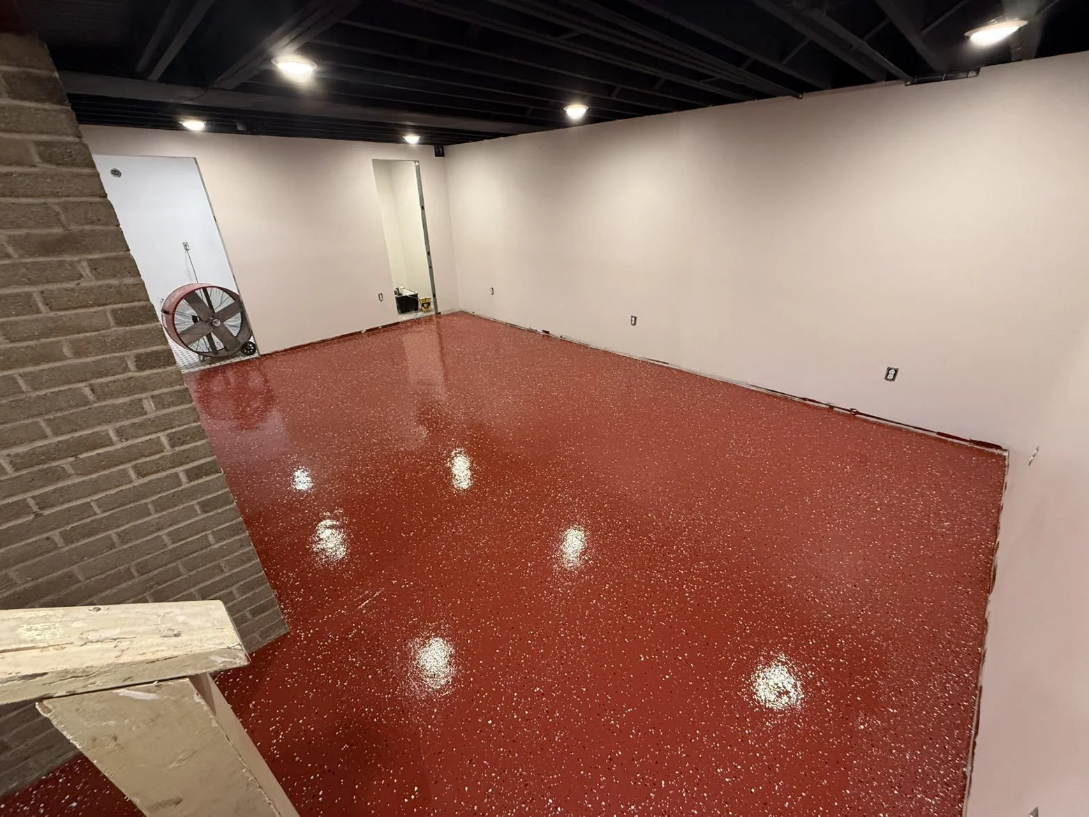 An empty room with a red polished concrete floor, white walls, and a black ceiling with recessed lighting. There is a door on the left wall and a large fan near the door.