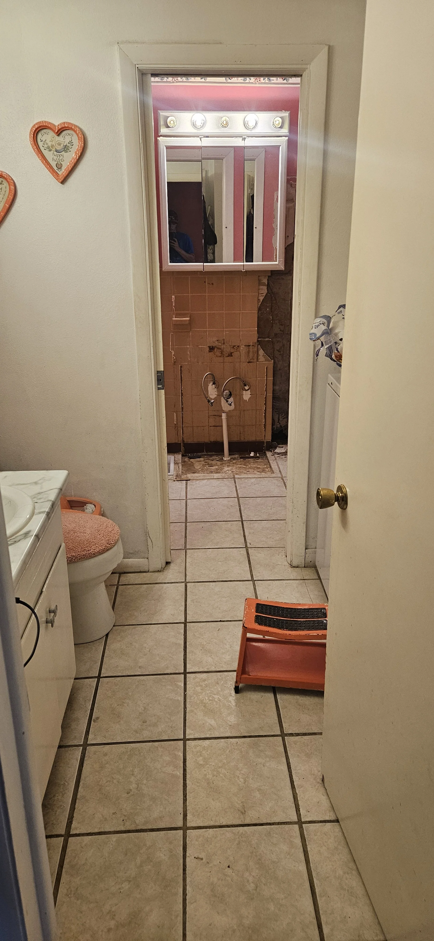 Bathroom with tiles, a white toilet with a pink lid cover, a small step stool, and a doorway leading to a room with exposed pipes and partially removed wall tiles.