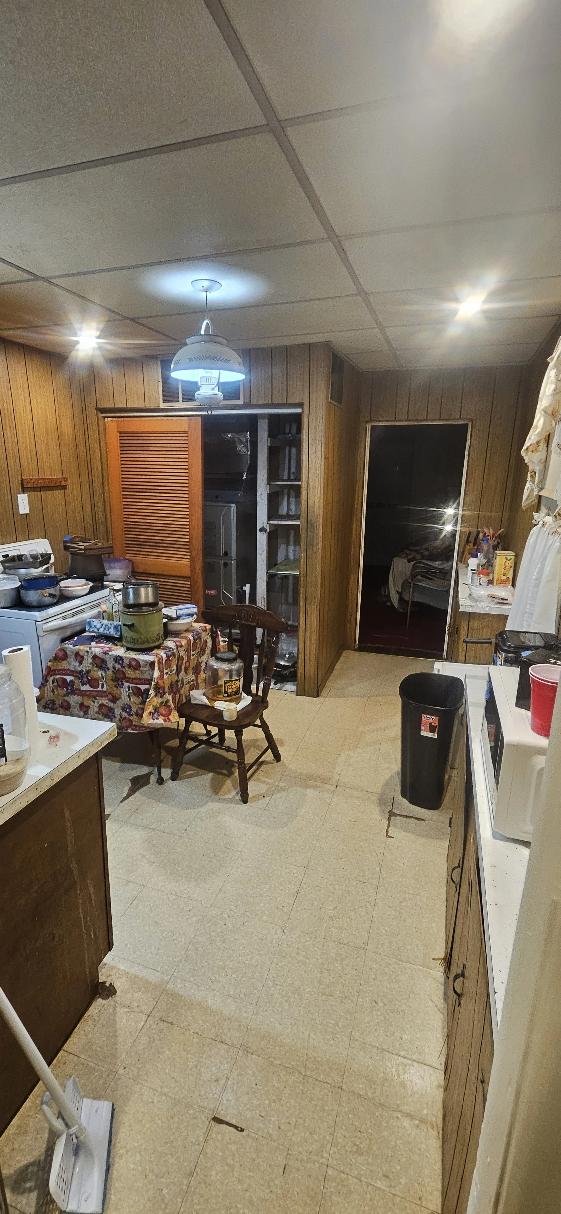 A cluttered kitchen with wooden paneling, a stove with pots, a small table with a tablecloth, and various kitchen items, leading to a doorway into a dark room.