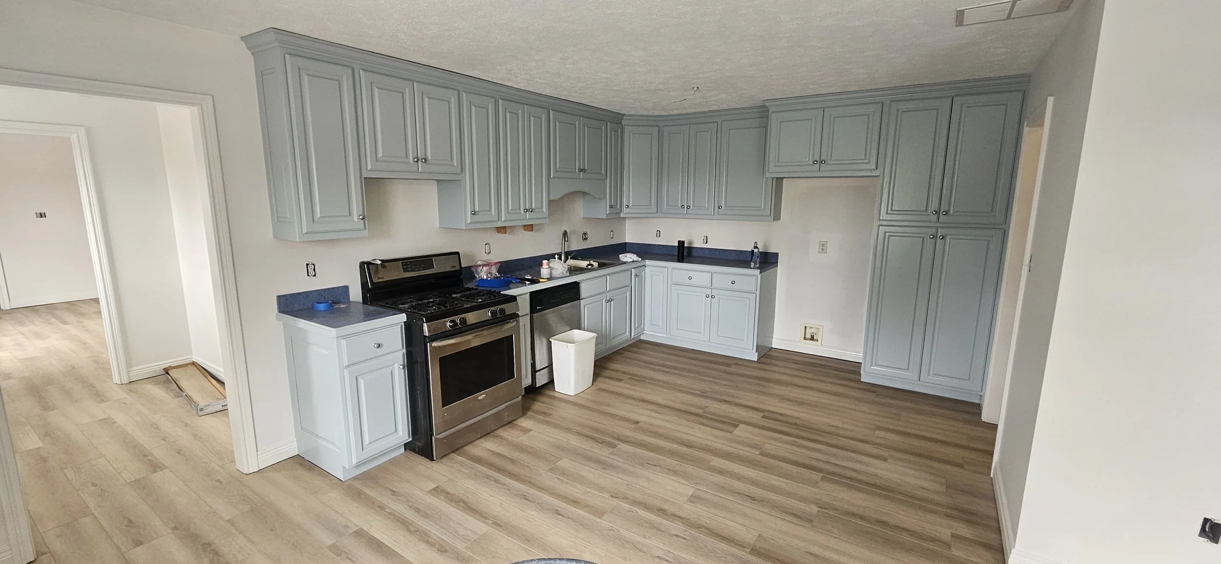 Empty kitchen with light blue cabinets, a stainless steel stove, and a black dishwasher, with a wooden floor and some construction materials on the floor.