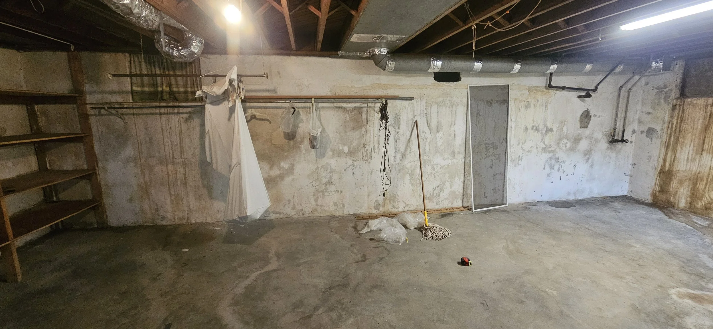 Empty basement or garage with unfinished walls, exposed pipes, shelving on the left, and cleaning supplies like mop, broom, and plastic bags on the floor.
