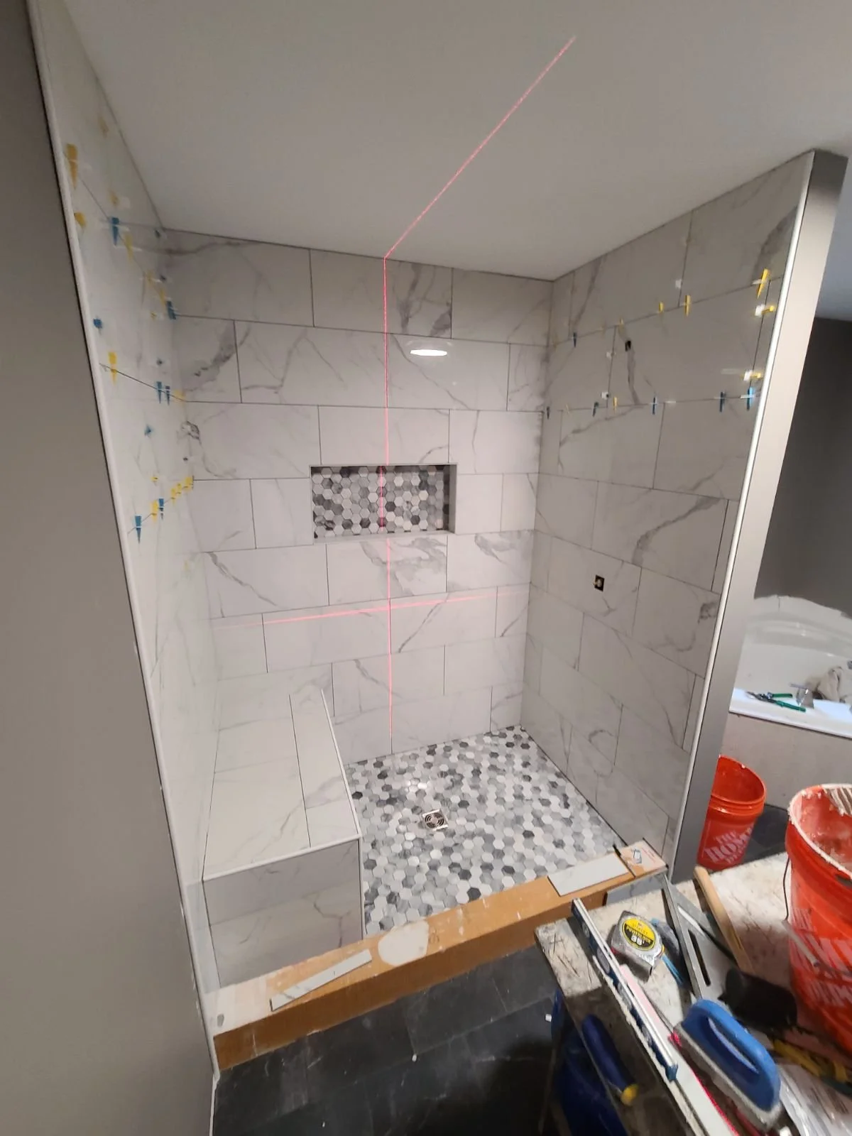 A bathroom shower area under construction with white marble tiles, a built-in bench, and a wall niche with hexagonal tiles, laser level and construction tools visible.