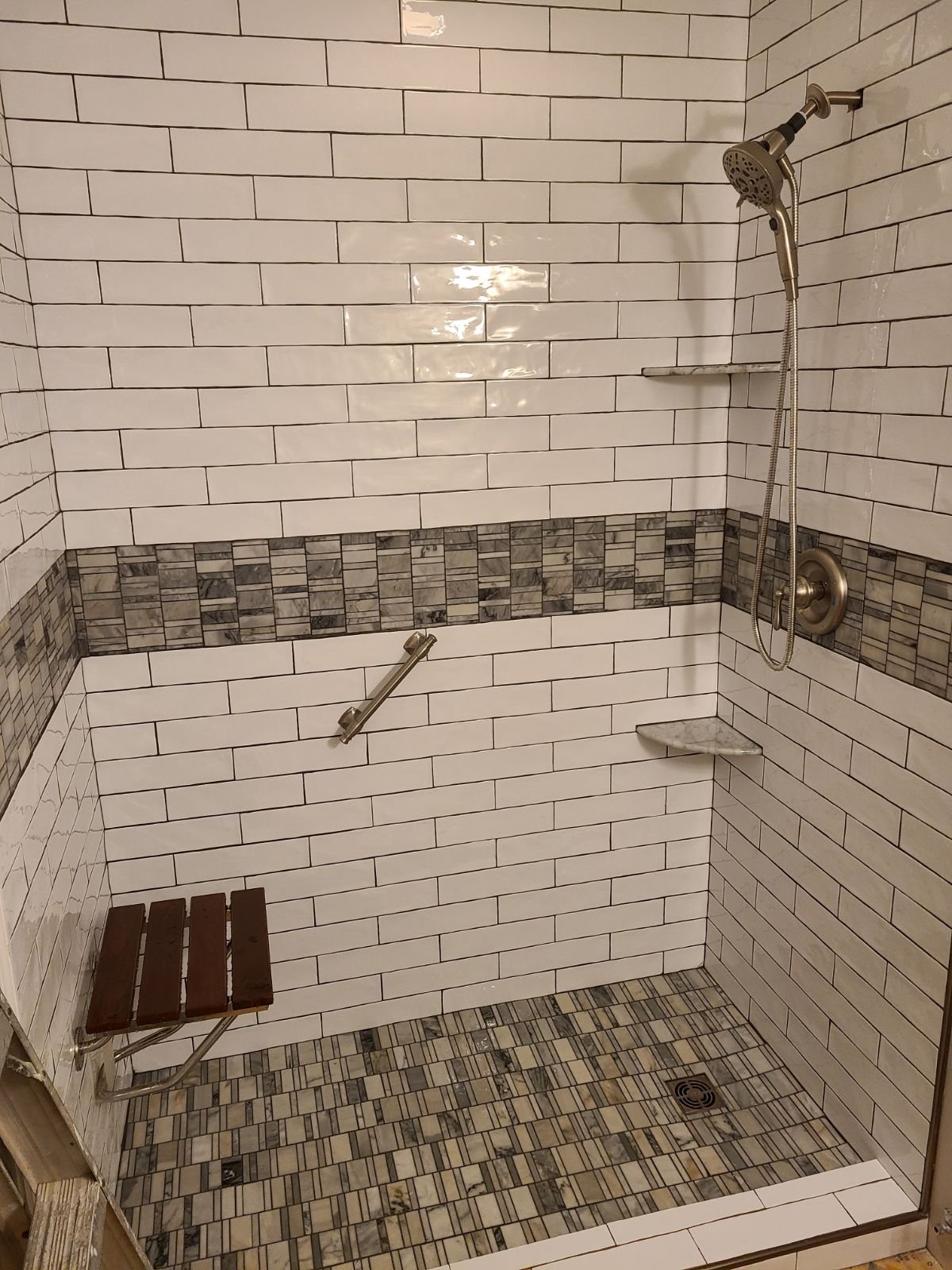 A tiled shower with white subway tiles, a horizontal strip of small gray stones, a handrail, a wooden bench, and marble shelves.