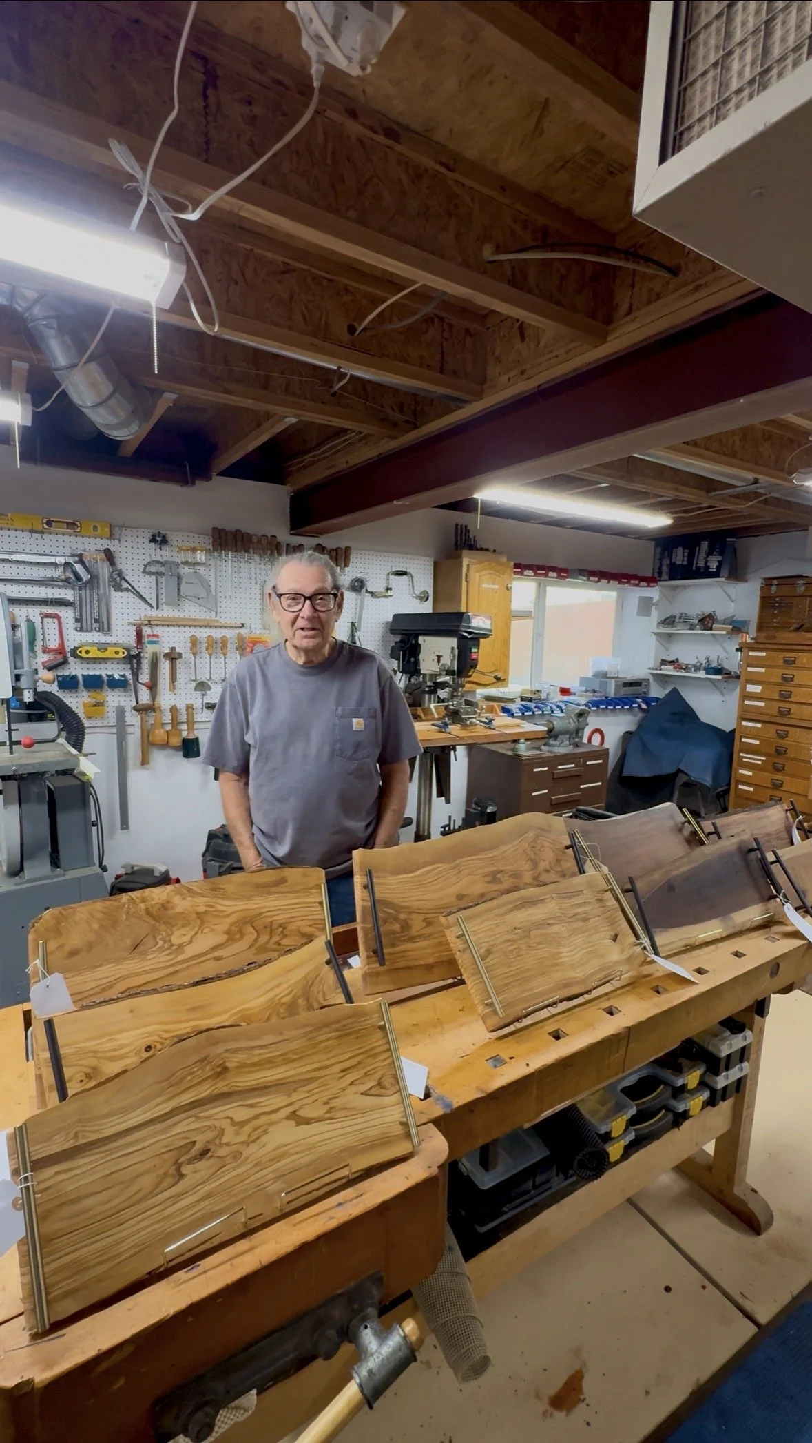An elderly man standing in a woodworking workshop with wooden furniture pieces and tools on the wall behind him.