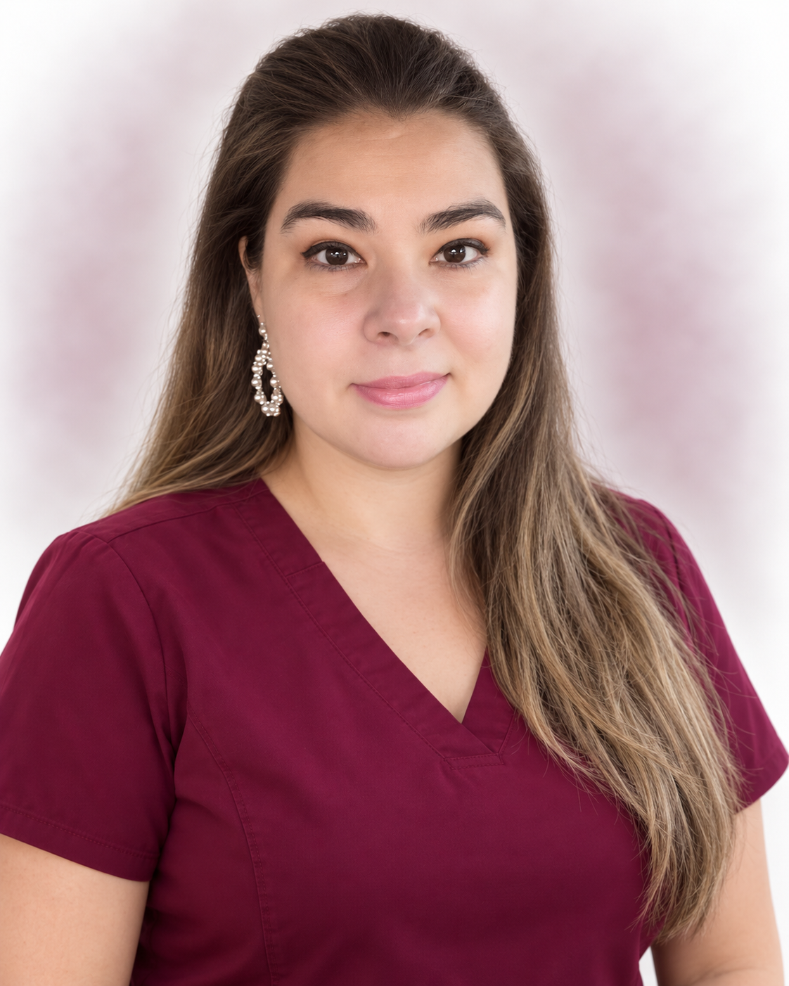 A woman with long brown hair wearing a burgundy V-neck top and decorative earrings, standing against a soft-focus background.