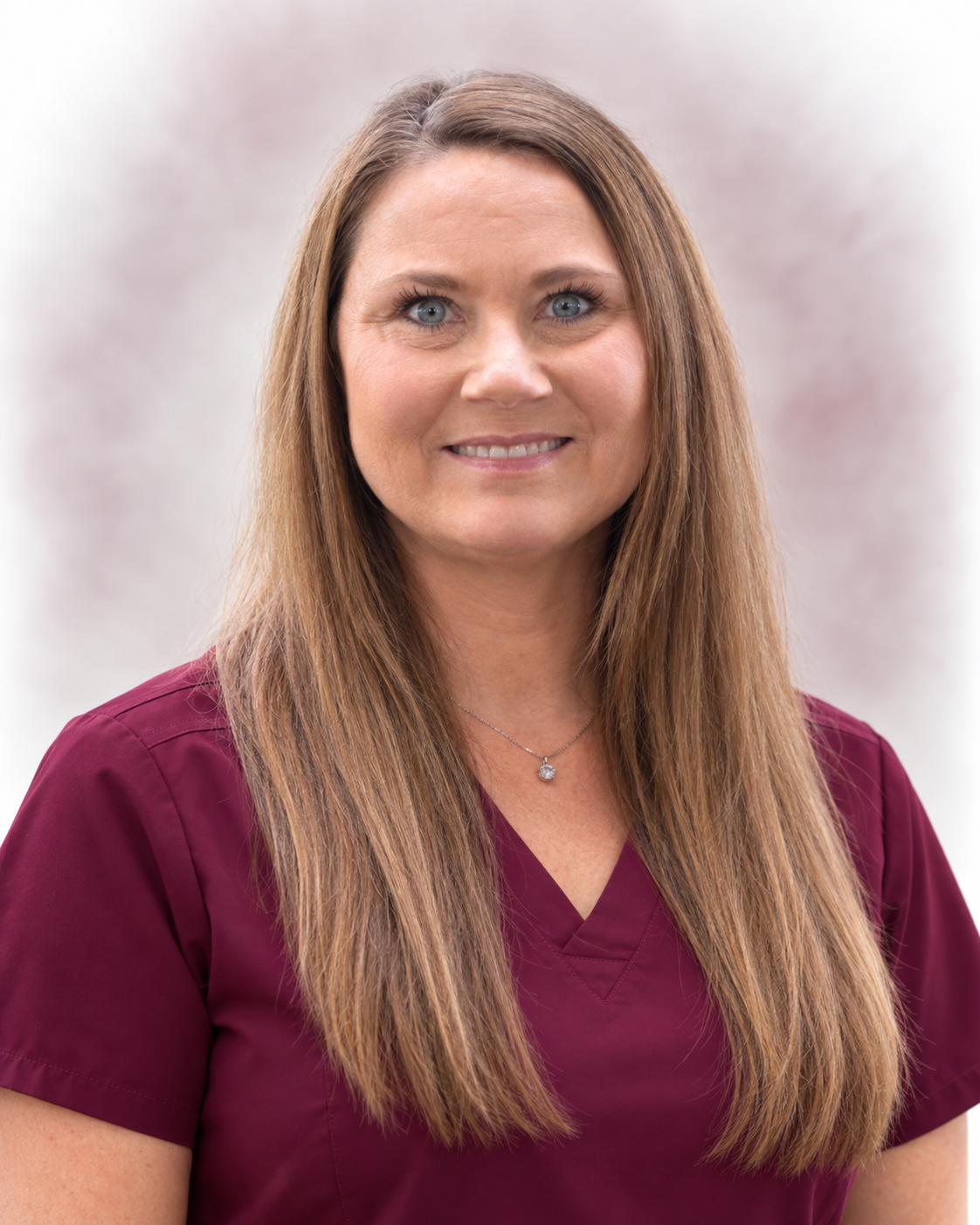 A woman with long light brown hair, blue eyes, and a friendly smile, wearing a maroon medical uniform and a silver necklace, posing against a plain light background.