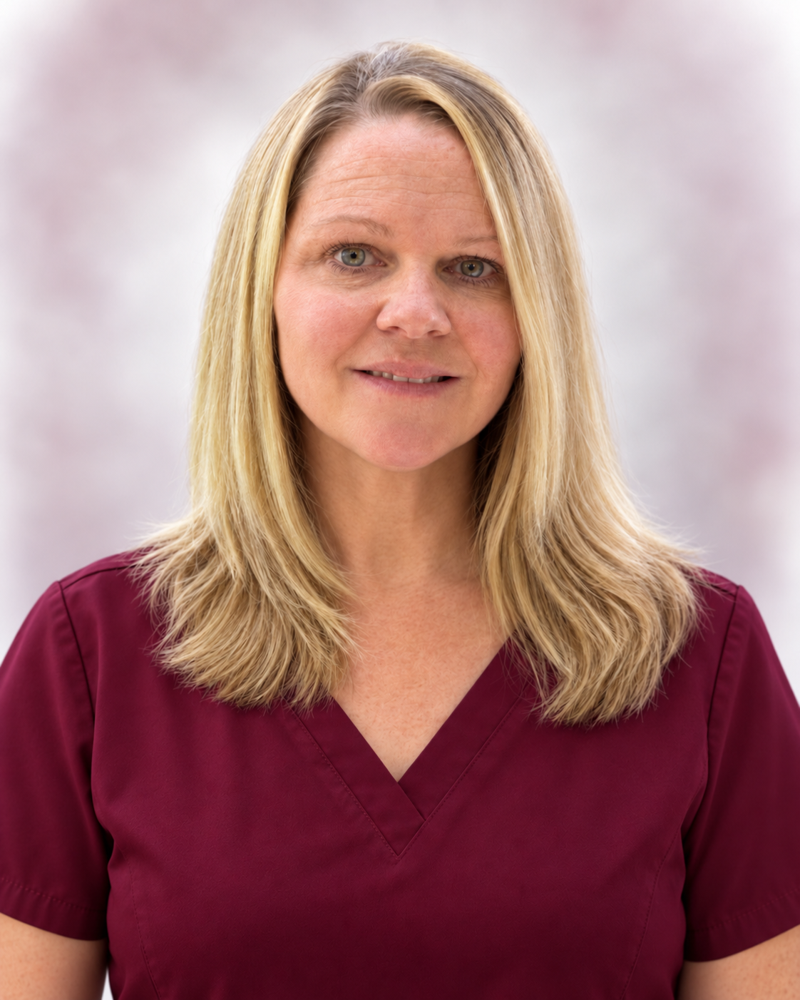 A woman with blonde hair wearing a maroon medical scrub top, smiling softly, standing against a blurred light background.