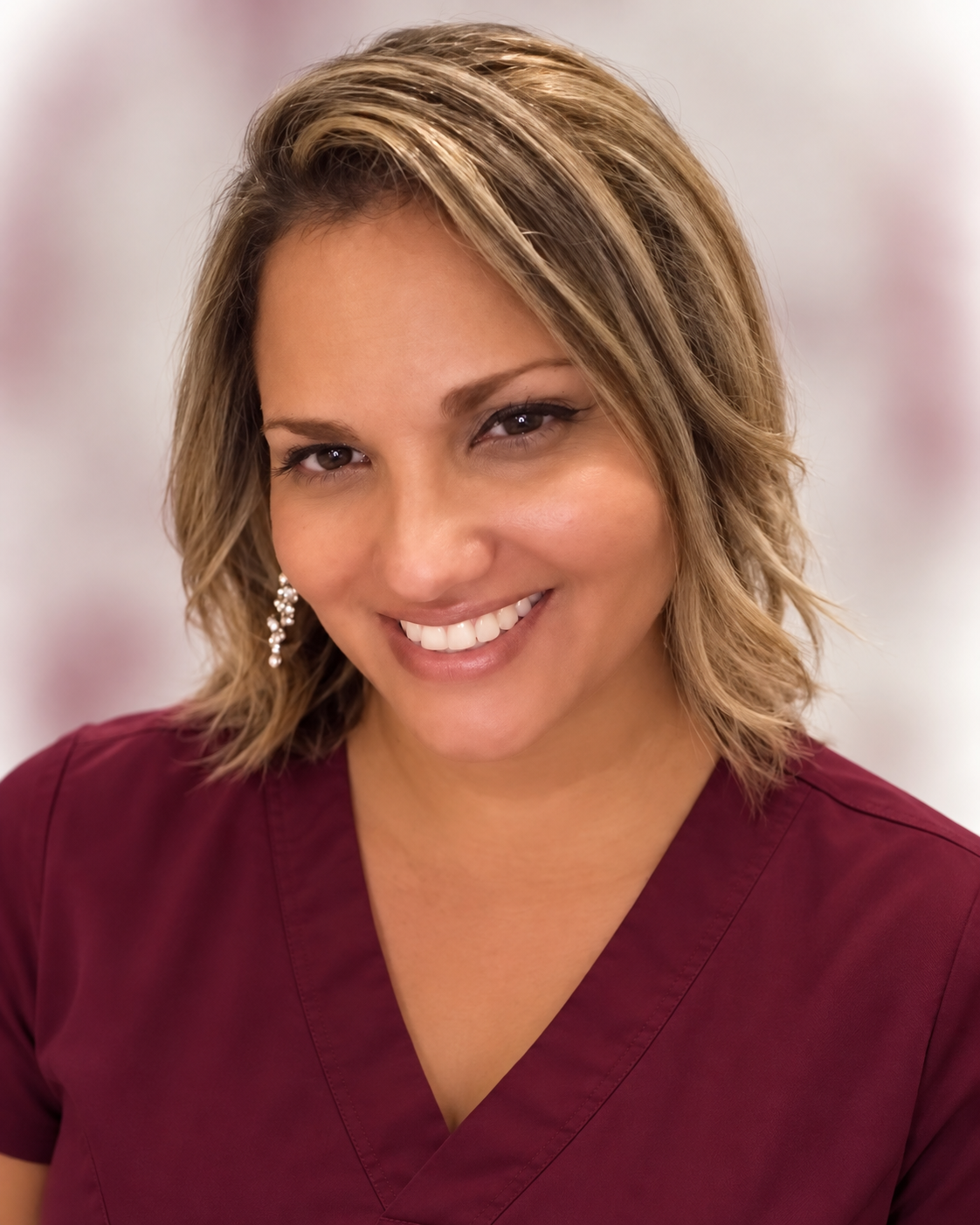 Close-up of a smiling woman with short, wavy blonde hair wearing a maroon top and pearl earrings.