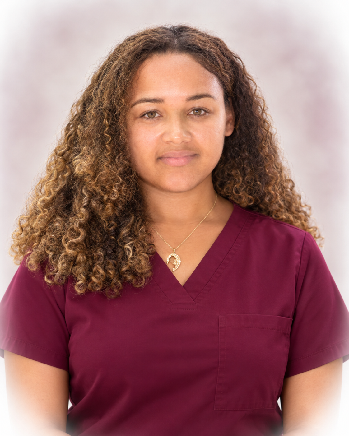 A woman wearing maroon medical scrubs with curly brown hair and a gold necklace with a moon pendant.