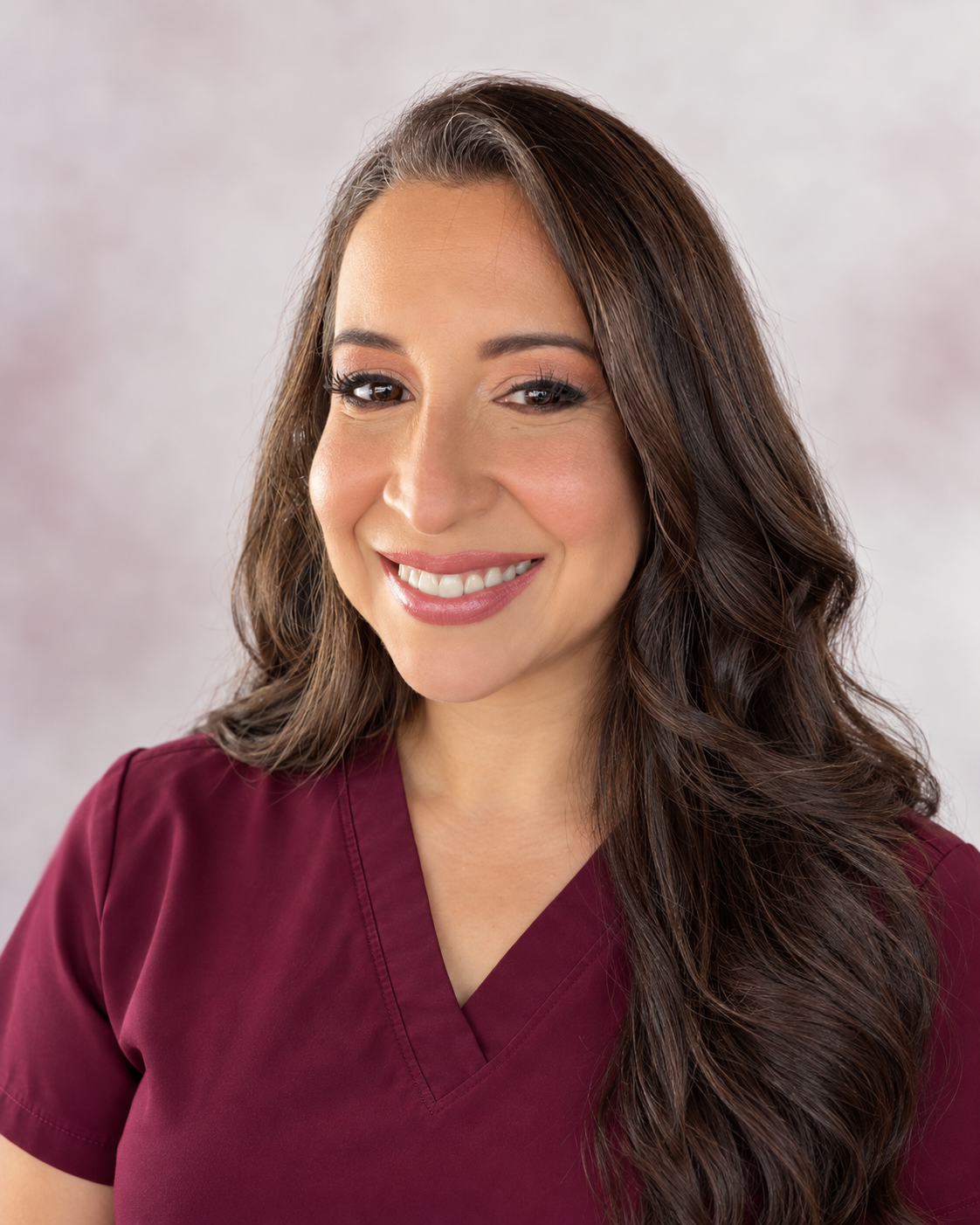 Headshot of a woman with long brown hair and brown eyes, smiling, wearing maroon scrubs, against a neutral background.