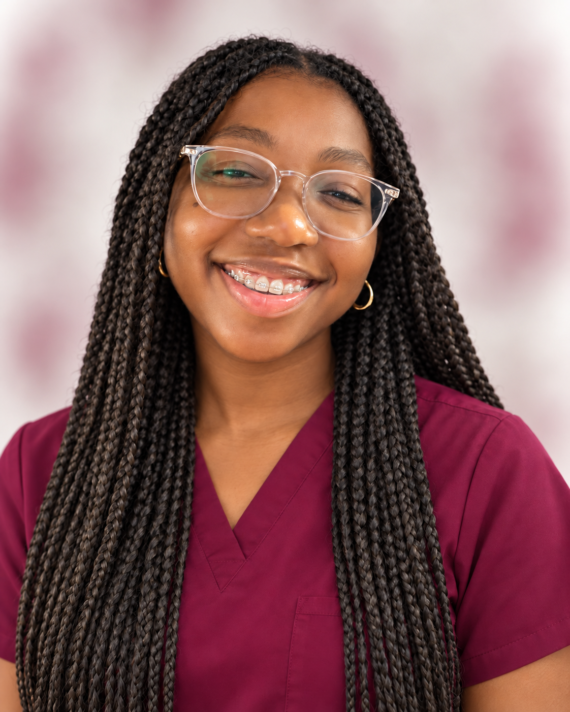 A young woman with long braided hair, wearing glasses and braces, smiling at the camera, dressed in maroon scrubs.