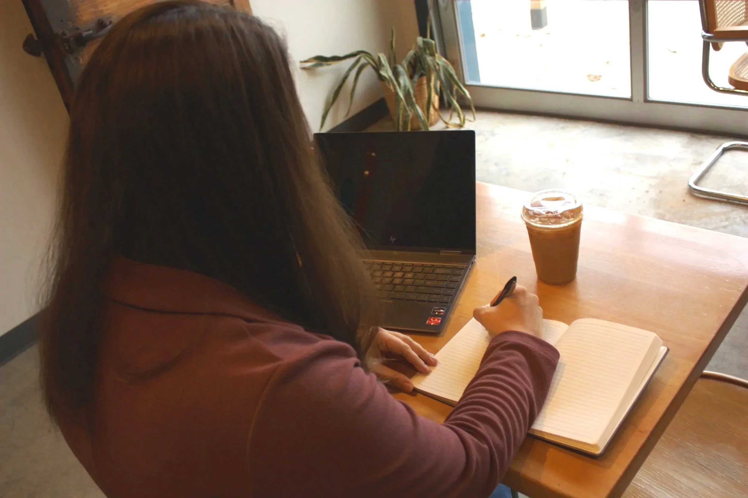A woman sitting at a wooden table, writing in a notebook, with a laptop, a pen, a cup of iced coffee, and a plant nearby, near a glass door letting in natural light.