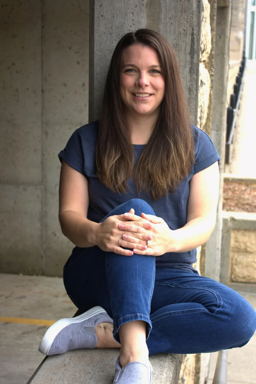 A woman with long brown hair sitting on a stone surface outdoors, leaning against a concrete wall, smiling at the camera, wearing a blue shirt, blue jeans, and gray sneakers.