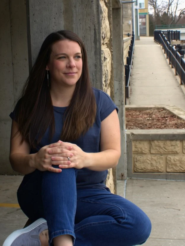 Woman sitting against a stone wall outside, wearing a navy T-shirt and jeans, with long brown hair and earrings.