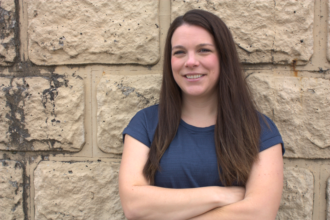 A woman with long brown hair, wearing a navy blue t-shirt, standing with her arms crossed in front of a beige brick wall, smiling.