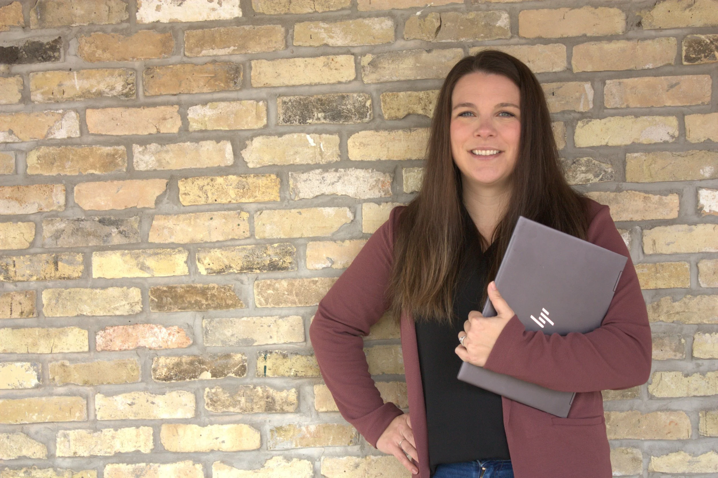 A woman with long brown hair wearing a burgundy blazer and black top holding a closed laptop with an HP logo, standing in front of a brick wall and smiling.
