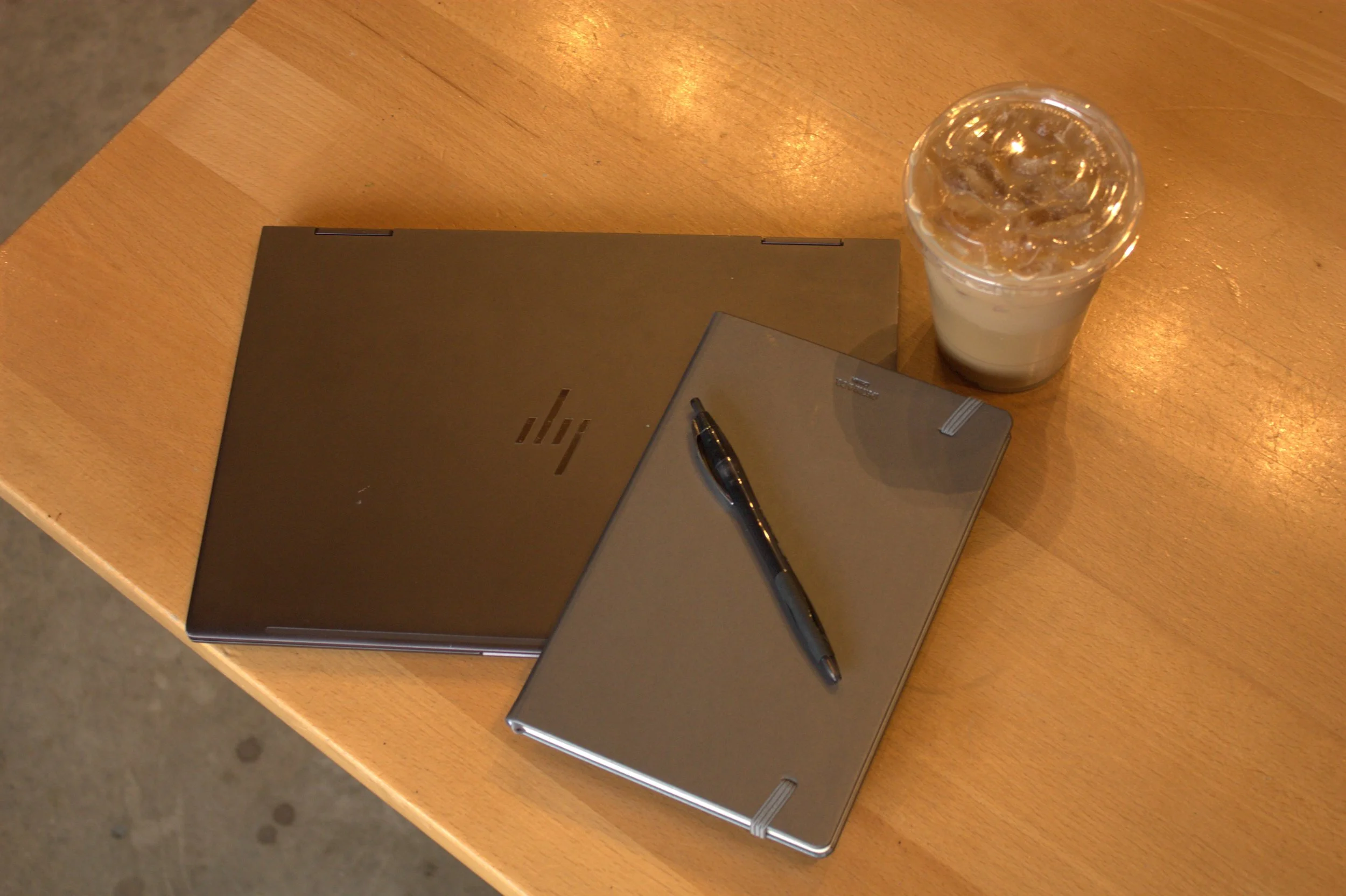 A wooden table with a closed black HP laptop, a black ballpoint pen, a smartphone, and a plastic cup filled with ice and a drink.