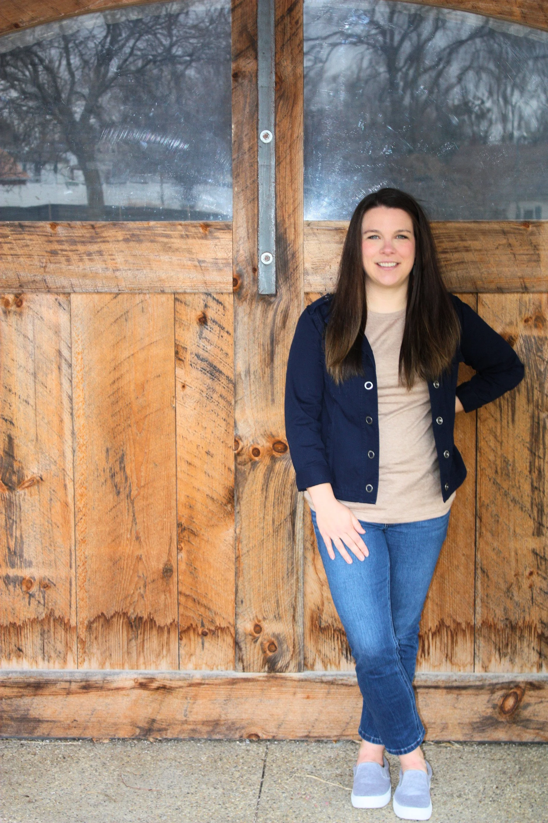 A woman with long dark hair wearing a beige shirt, blue jacket, and jeans standing in front of a weathered wooden barn door with glass windows, smiling with her hand on her hip, in an outdoor setting.