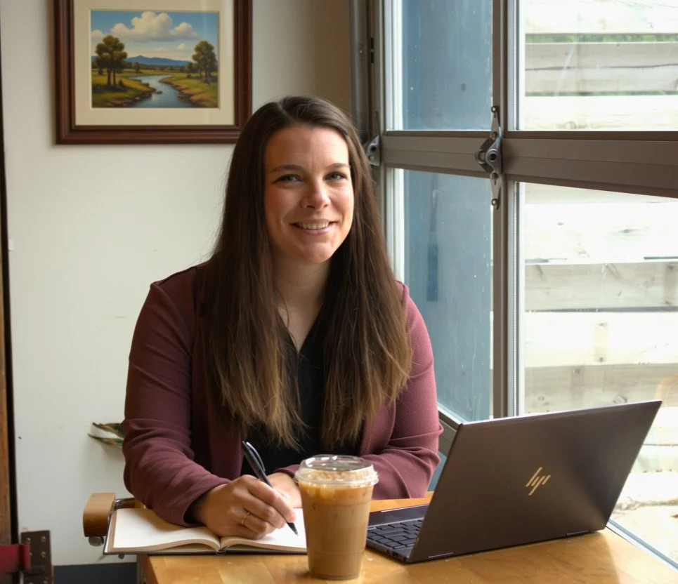 A woman with long brown hair smiling at a table with a laptop and iced coffee, sitting near a window with a landscape painting on the wall behind her.