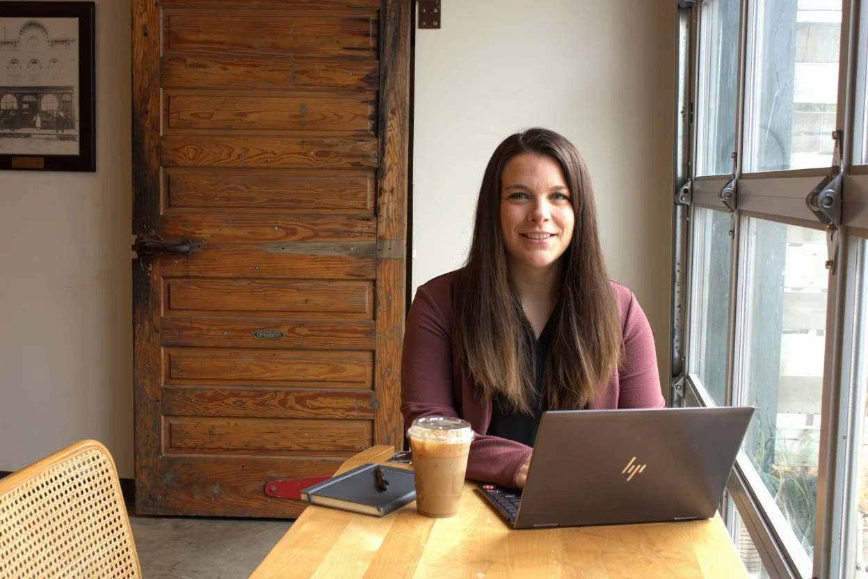 A woman with long brown hair sitting at a wooden table with a laptop, an iced coffee, a notebook, and a pen in a bright room near large windows and a wooden door.