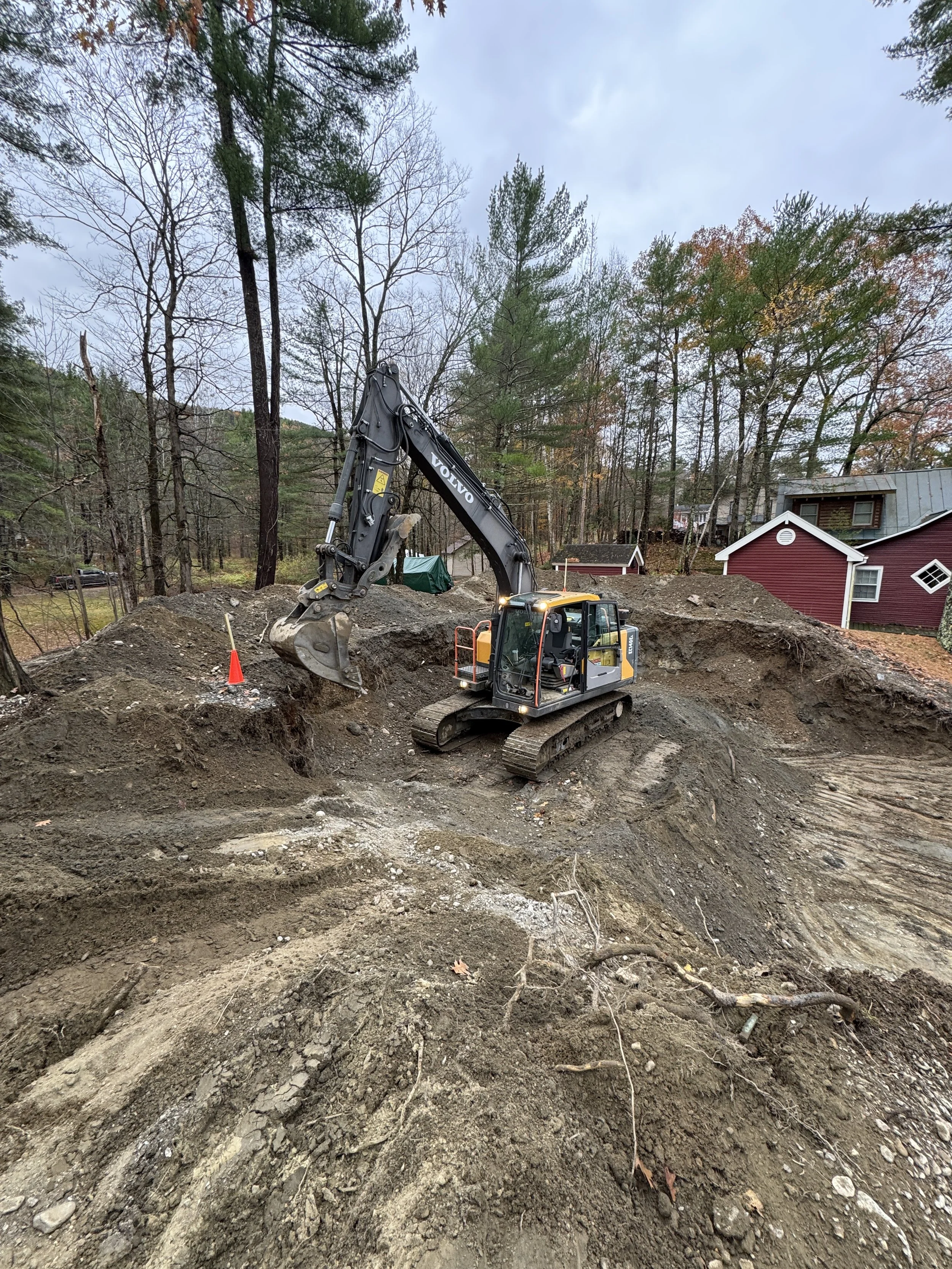 Construction site with a yellow excavator digging in the dirt near trees and some residential houses in the background.