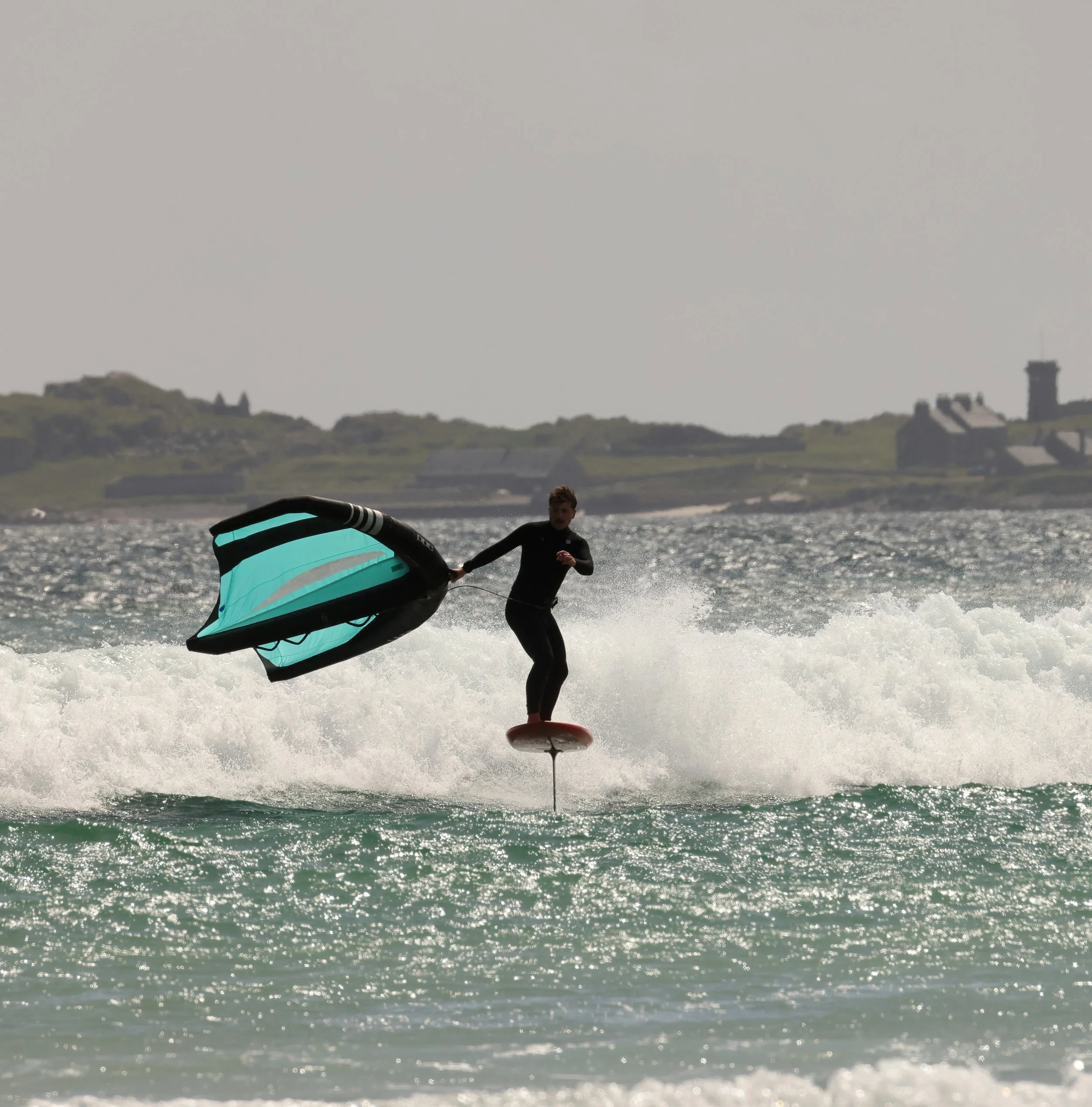 A person surfing on a wave in the ocean, holding onto a kite with a bright turquoise sail, with houses and a hill in the background under a gray sky.