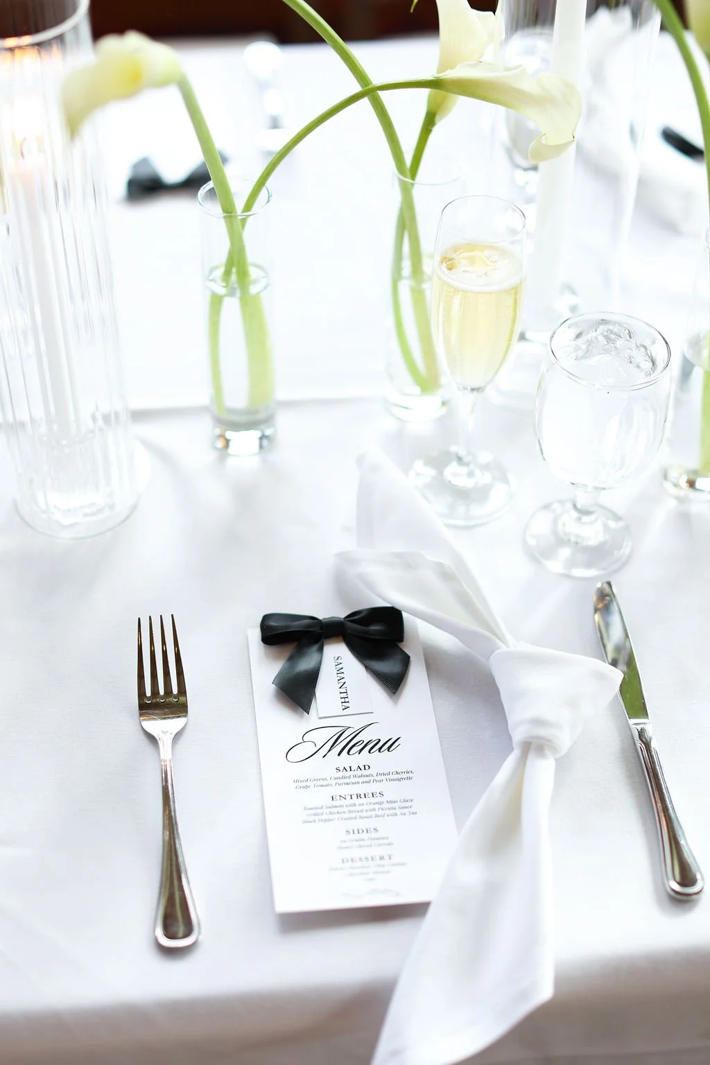 Elegant table setting with white tablecloth, silverware, a menu with a black bow, white napkin, glassware, and vases with white calla lilies and water. A champagne flute and a water glass with ice are also visible.