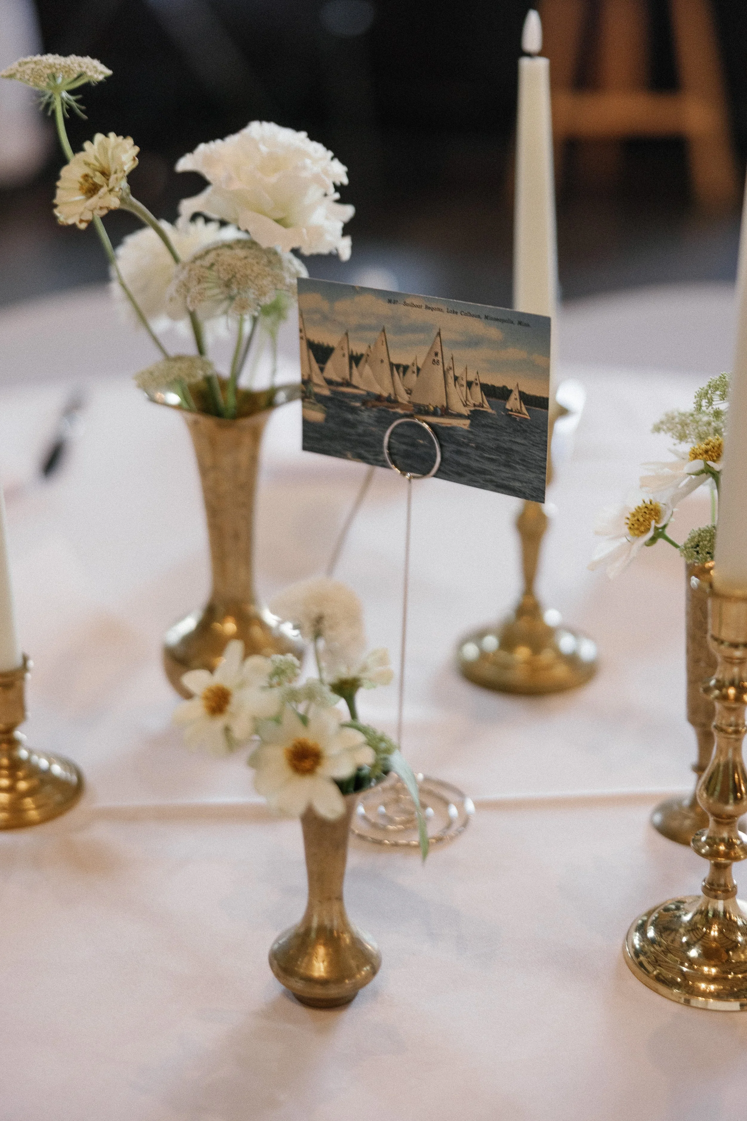 Decorative table centerpiece with white flowers in gold vases and a photograph of sailboats on the water, set on a white tablecloth.