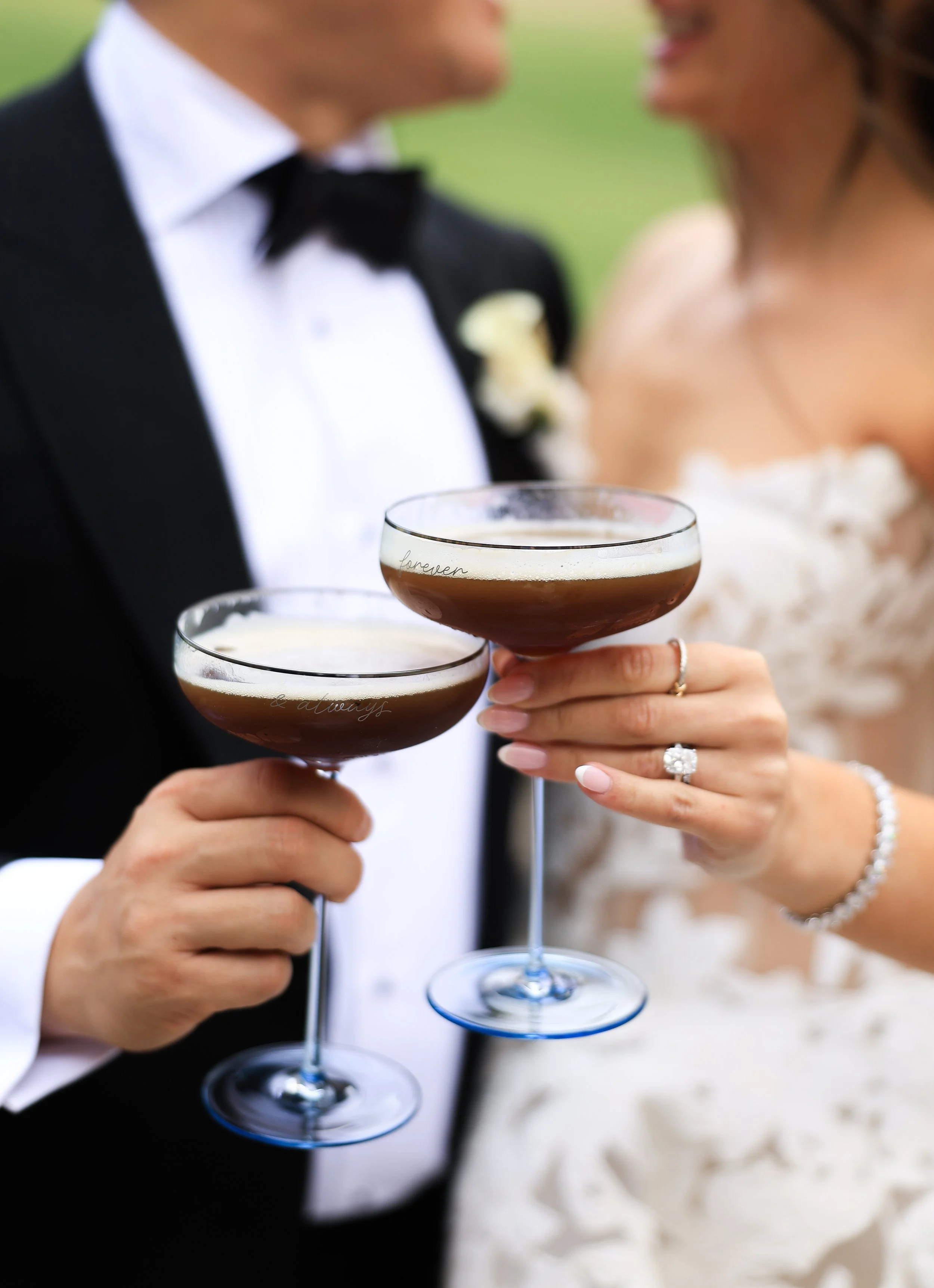 Close-up of a newlywed couple holding champagne glasses, with blurred background of a bride in a wedding dress and groom in a tuxedo.