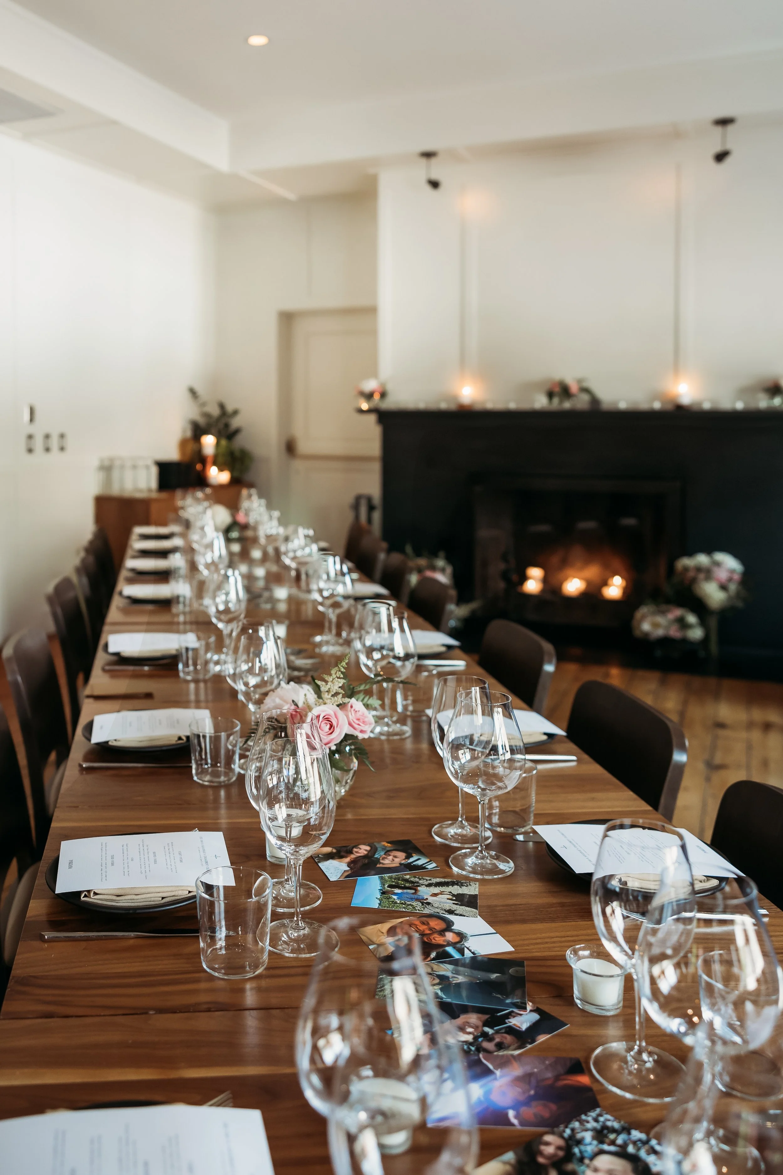 A long wooden table set for a meal with glasses, plates, and floral centerpieces in a cozy indoor setting with a fireplace and decorative candles.