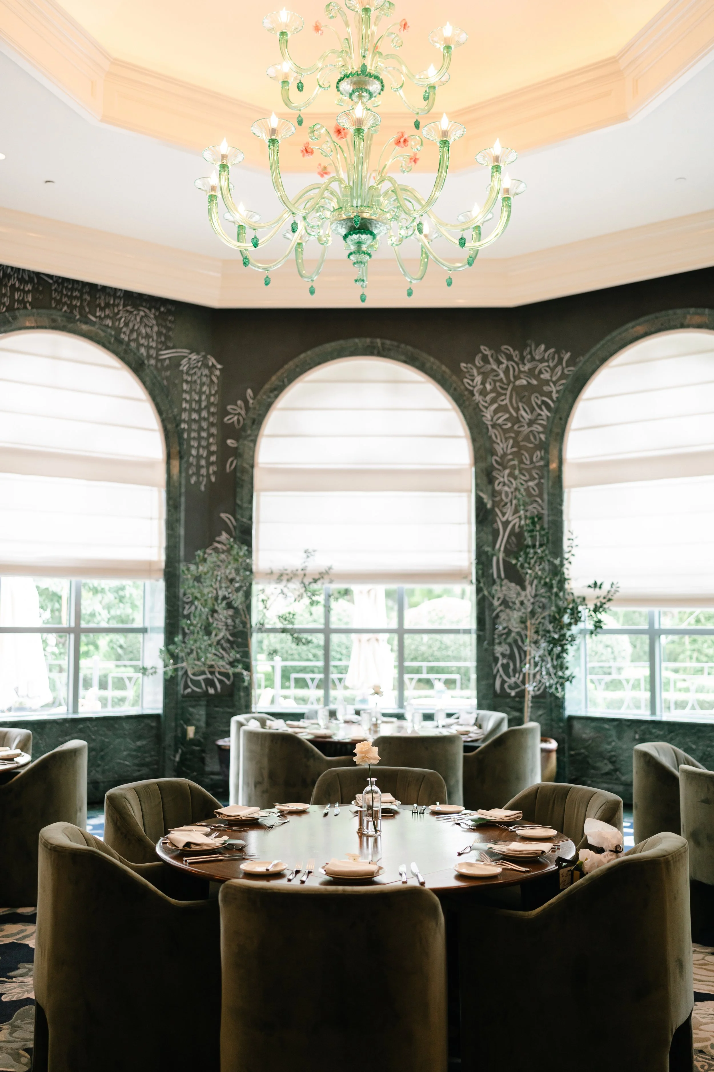 Elegant restaurant dining area with a round table set with napkins, plates, and silverware. Surrounding beige velvet chairs, large windows with white blinds, dark patterned walls with floral designs, and a large glass chandelier hanging from the ceiling.