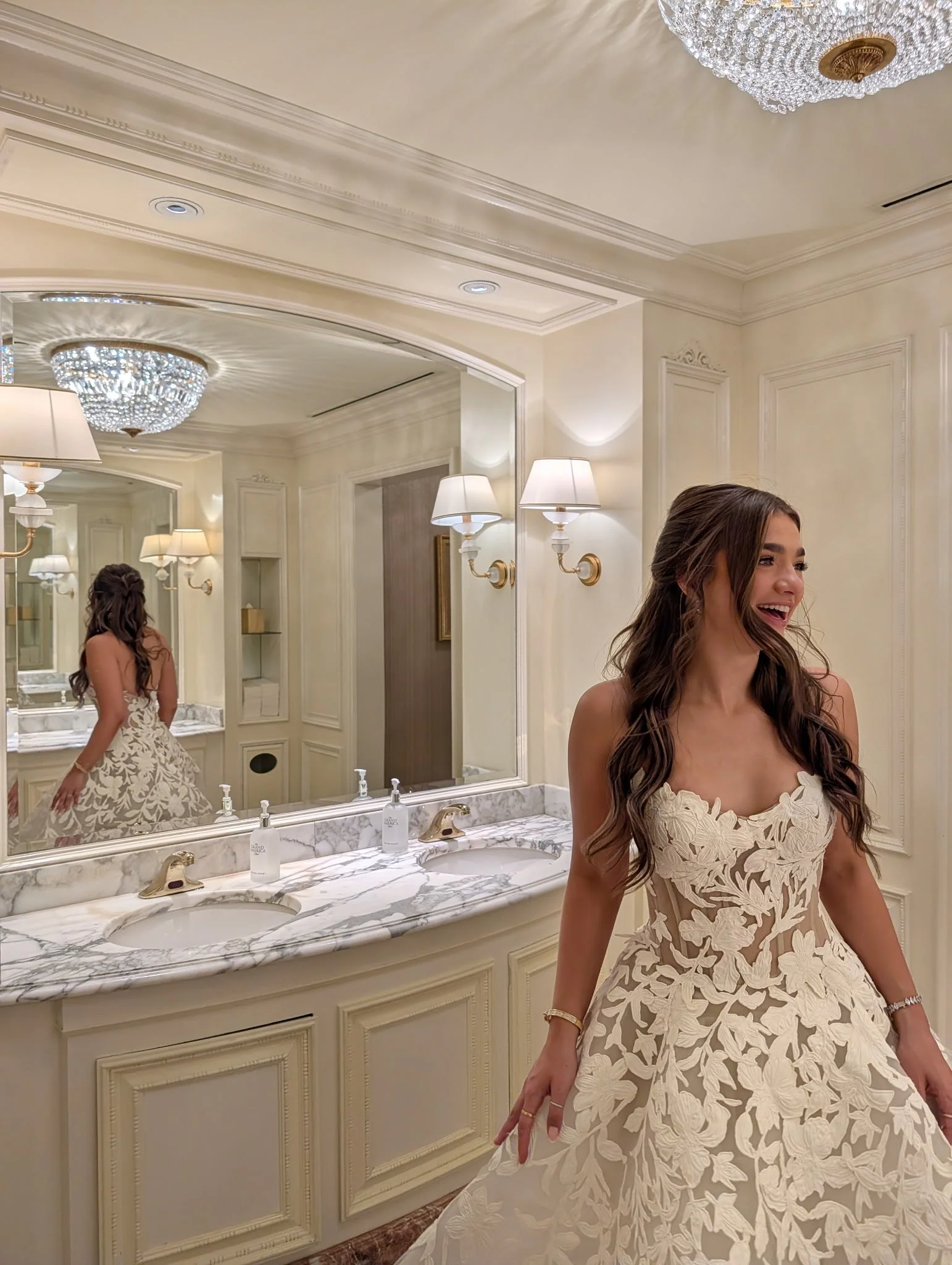 A woman in a white, floral lace dress standing in front of a bathroom mirror with a marble countertop and a chandelier overhead, smiling.