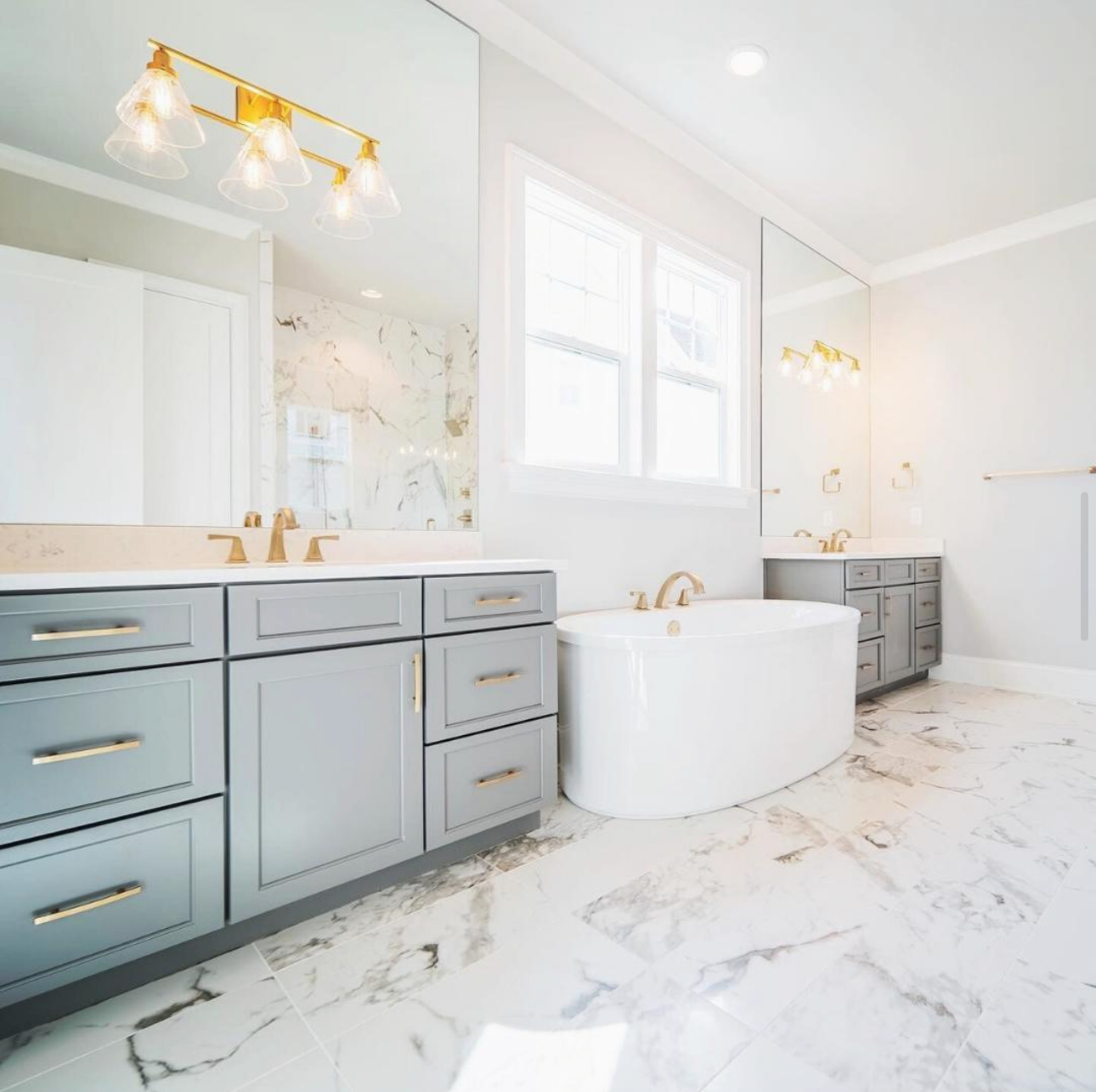 Bright modern bathroom with gray double vanity, gold fixtures, marble tile flooring, mirrored walls, and freestanding white bathtub